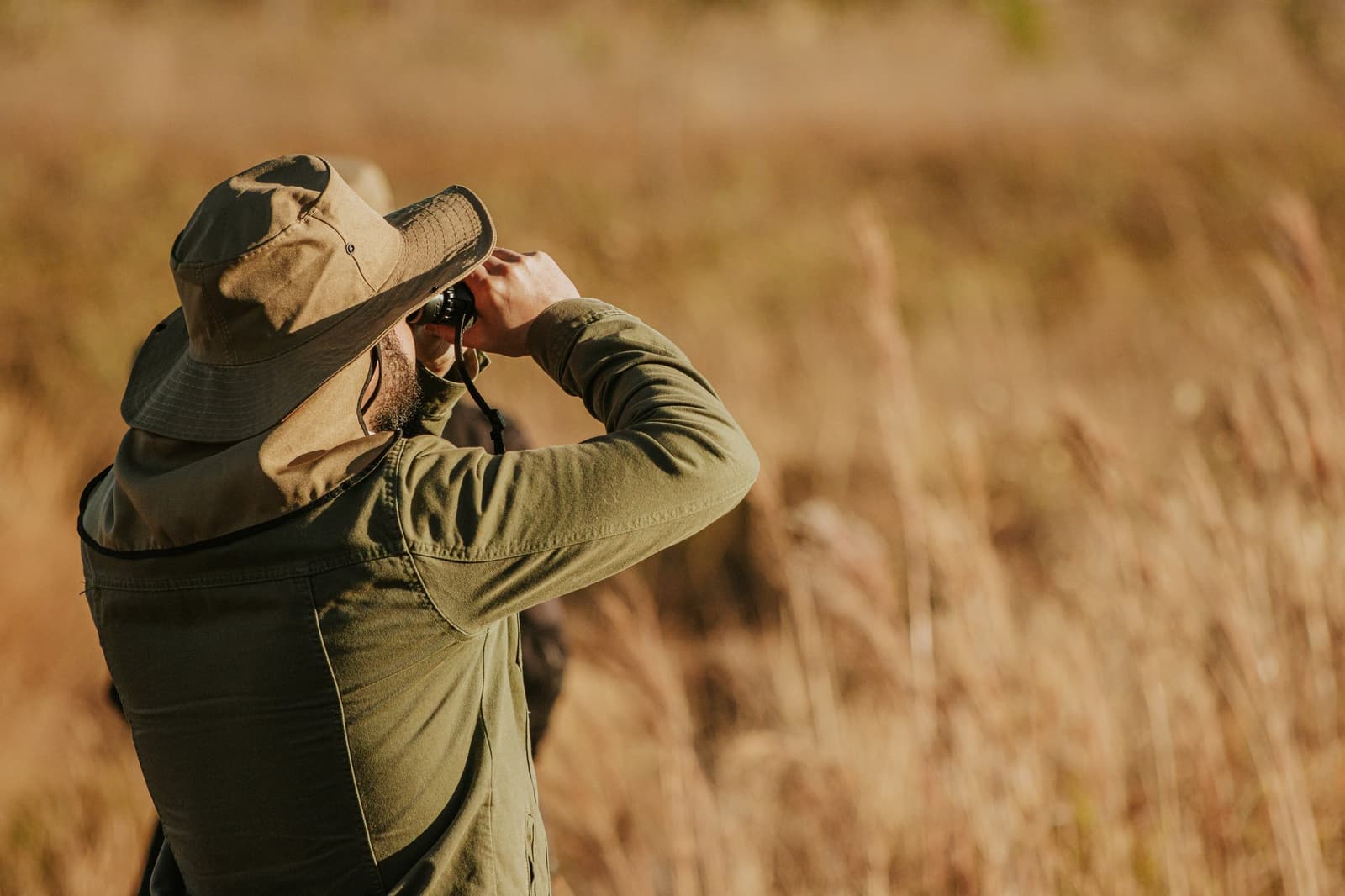 Dans le Cerrado, chaque bruissement dans les herbes vaut le coup d'œil. Un voyageur barbu coiffé d'un chapeau à large bord observe à travers des jumelles les hautes herbes dorées du Cerrado lors d'une sortie ornithologique à la Pousada Trijunção
