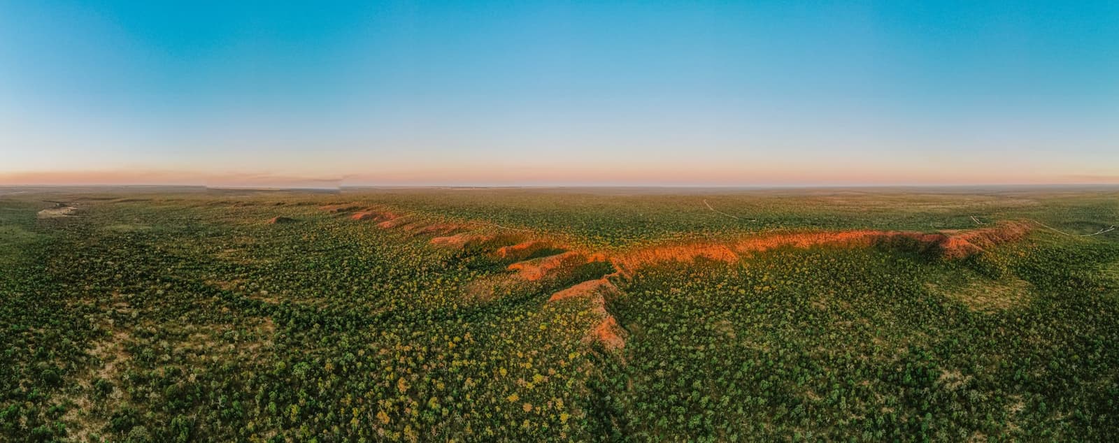 Le Cerrado s'étend jusqu'à l'horizon, cette bande rouge sang rappelant que la savane a son propre caractère. Vue aérienne panoramique du paysage de Cerrado autour de la Pousada Trijunção au coucher du soleil, avec une crête de latérite rouge vif traversant une savane verdoyante sous un ciel rosé et bleu pastel