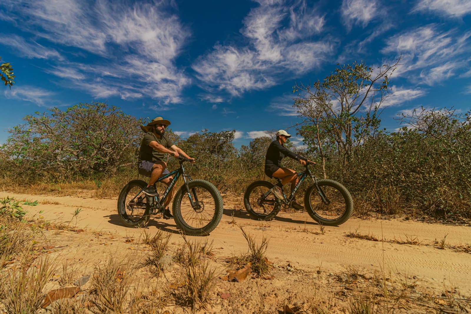 Le sable, le vent sec et deux vélos qui tiennent bon. Le Cerrado a ses propres règles. Deux cyclistes sur des fat bikes longeant un sentier sablonneux du Cerrado à Pousada Trijunção, entourés d'arbustes natifs sous un ciel bleu intense