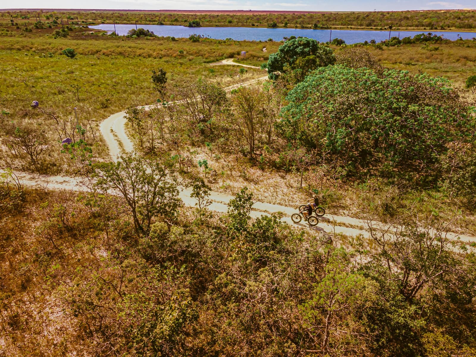 Le chemin se faufile entre les arbustes, et le lac attend au bout. Vue aérienne de deux cyclistes sur fat bikes longeant un sentier de terre sinueux à travers la végétation du Cerrado à la Pousada Trijunção, avec une lagune bleue visible en arrière-plan