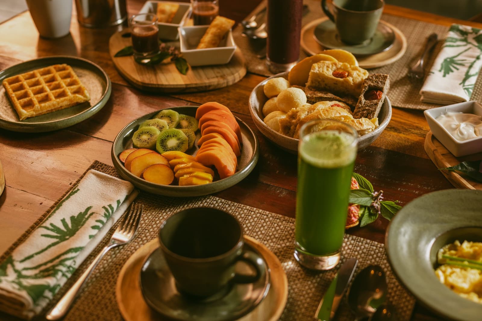 Un petit-déjeuner qui donne envie de prendre son temps, même quand la faune n'attend pas. Table de petit-déjeuner généreuse à la Pousada Trijunção, garnie de tranches de kiwi, pêche et papaye, de gaufres dorées, de petits pains, d'un jus vert vif et d'une tasse de café en céramique