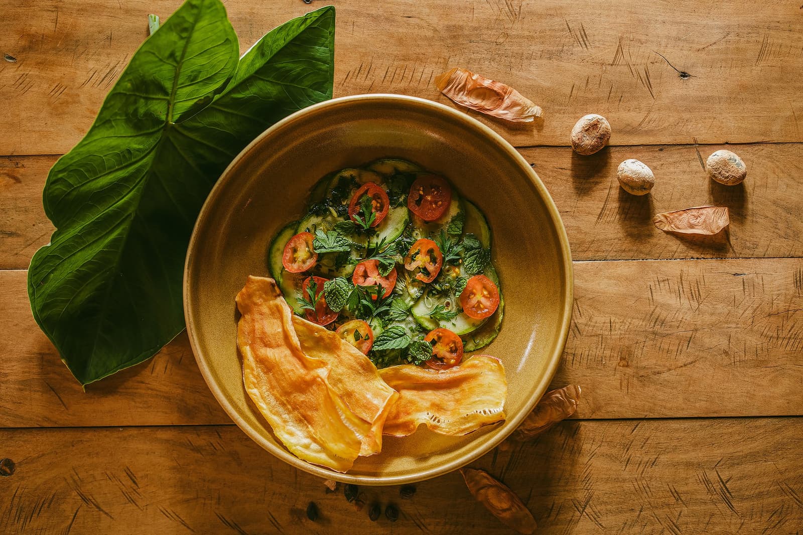 Un plat qui sent bon la forêt autant qu'il la respecte. Vue de dessus d'un bol en céramique garni de courgettes, tomates cerises et herbes fraîches accompagnés de chips croustillants à la Pousada Trijunção, entouré de feuilles tropicales et de graines sauvages sur une table en bois rustique