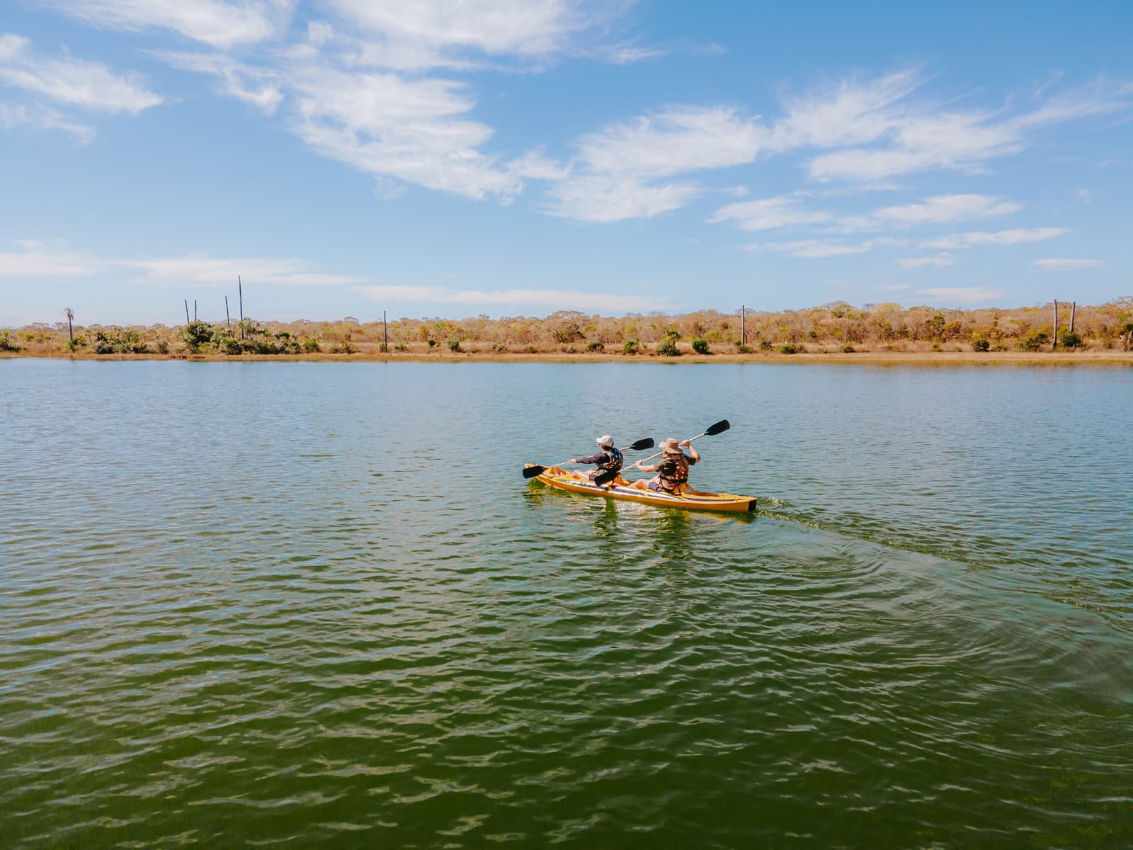 En kayak, le Cerrado ralentit. On remarque enfin tout ce qu'on aurait manqué à pied. Deux pagayeurs dans un kayak tandem jaune glissant sur les eaux calmes et verdâtres de la Lagoa das Araras à la Pousada Trijunção, avec la forêt sèche du Cerrado visible sur la rive opposée sous un ciel bleu ensoleillé