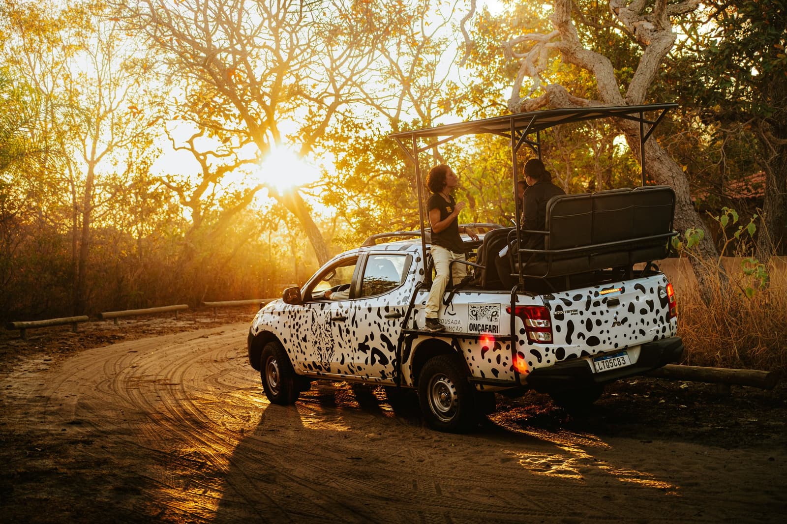 Le soleil se couche, la conversation, elle, ne s'arrête pas. Véhicule safari Onçafari aux taches de jaguar avec des visiteurs à bord arrêté sur une piste de terre au coucher de soleil doré dans le Cerrado de la Pousada Trijunção