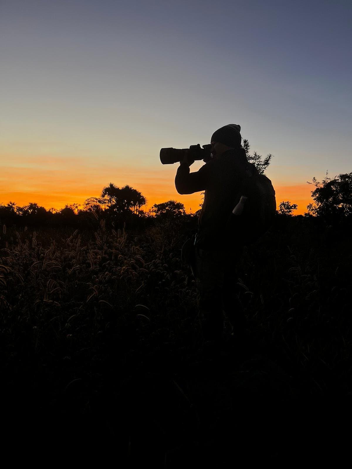 Quelques minutes de lumière, pas une de perdue. Silhouette d'un photographe animalier avec un téléobjectif levé vers le ciel orange et bleu de l'aube dans les hautes herbes du Cerrado à la Pousada Trijunção