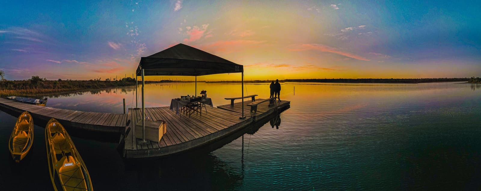 Dîner au bout du ponton, le ciel du Cerrado rien que pour vous. Silhouette d'un couple au bout d'un ponton en bois de la Pousada Trijunção, une table de petit-déjeuner sous auvent dans leur dos et des kayaks amarrés à gauche, sous un lever de soleil doré se reflétant sur le lac