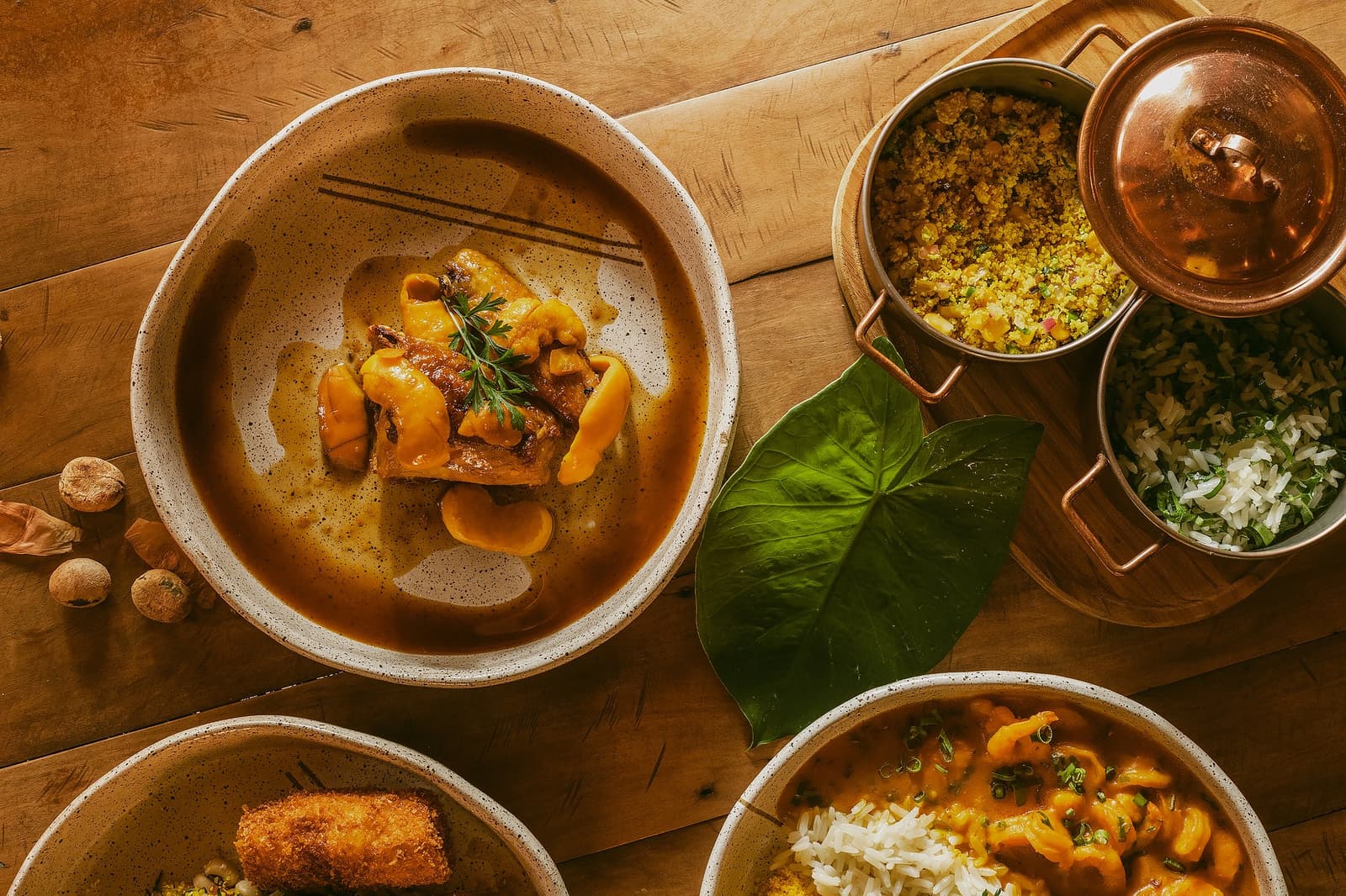 Un repas qui raconte le Cerrado mieux que n'importe quel guide. Vue de dessus d'un repas de la Pousada Trijunção comprenant un ragoût de viande braisée dans un bol en céramique mouchetée, une farofa dorée et du riz aux herbes dans des casseroles en cuivre, et un ragoût de fruits de mer crémeux avec du riz blanc, disposés sur une table en bois rustique