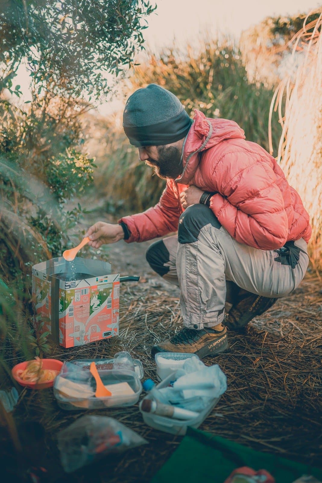 Dinner sorted, altitude and all. A bearded trekker in a red down jacket crouches over a portable camp stove on a dry grass clearing in the Serra da Mantiqueira, stirring a meal with an orange spoon during Serra Fina Trekking