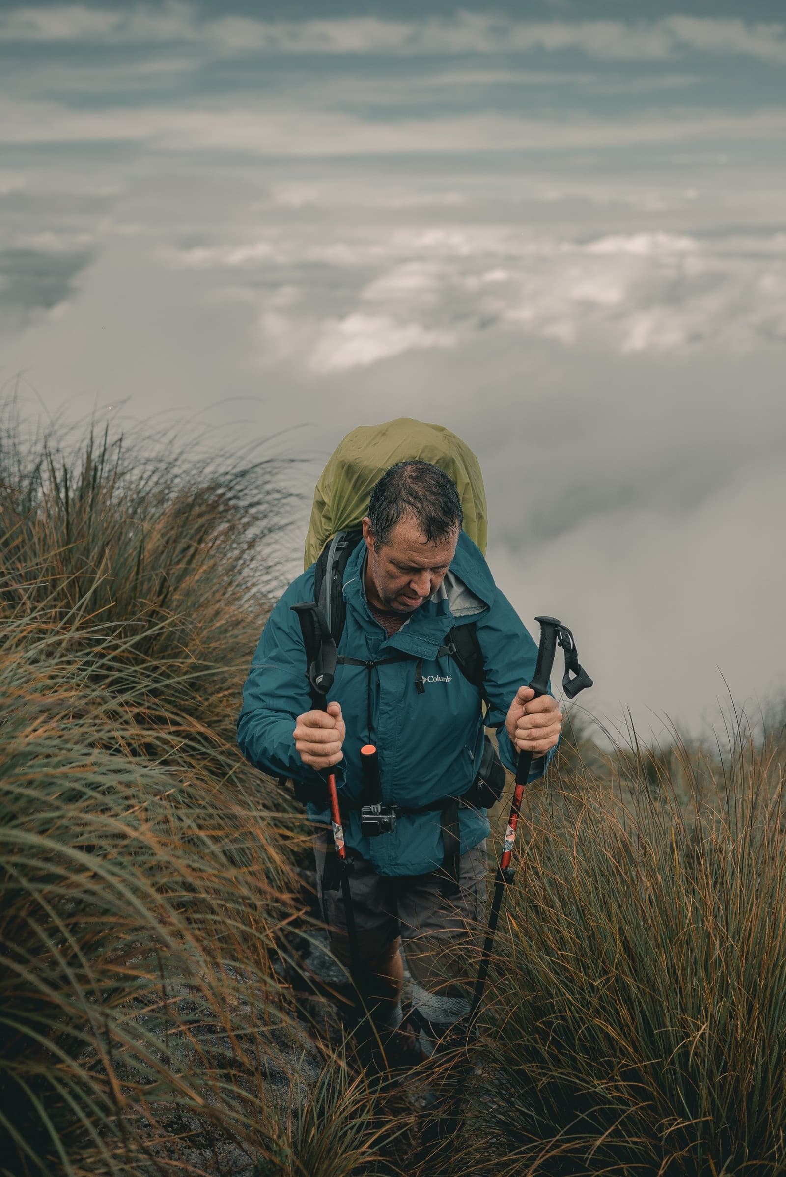 Some trails ask politely. This one just asks. A hiker carrying a full pack and trekking poles pushes through tall grass on a Serra Fina Trekking ridge, with a dramatic sea of clouds filling the valley far below