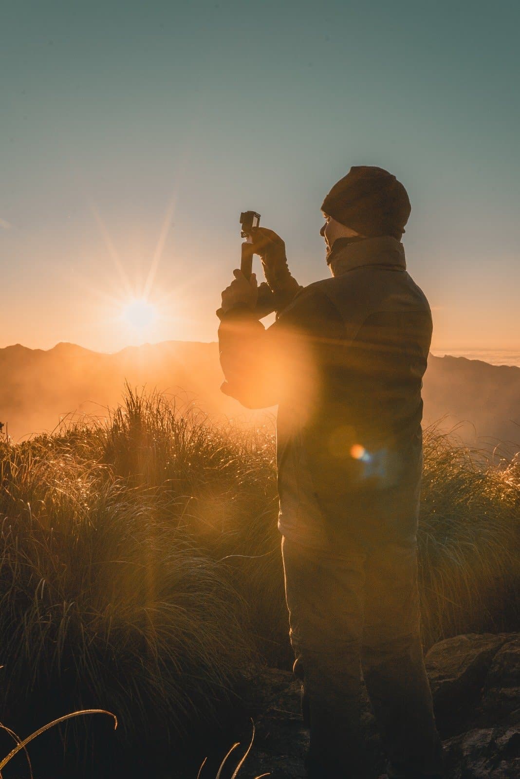 Up before the mountain wakes, camera already rolling. A hiker in a beanie and puffer jacket photographs the golden sunrise over misty Serra da Mantiqueira ridges during a Serra Fina trekking expedition, wild grasses glowing in the foreground