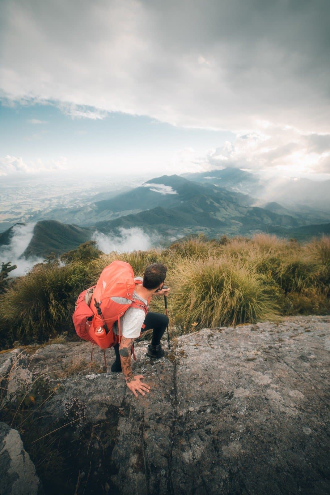 That moment at the top when the valley finally opens up below you. A hiker with a large red backpack and trekking pole crouches on a rocky ledge during Serra Fina Trekking, gazing over a vast misty mountain landscape in the Serra da Mantiqueira