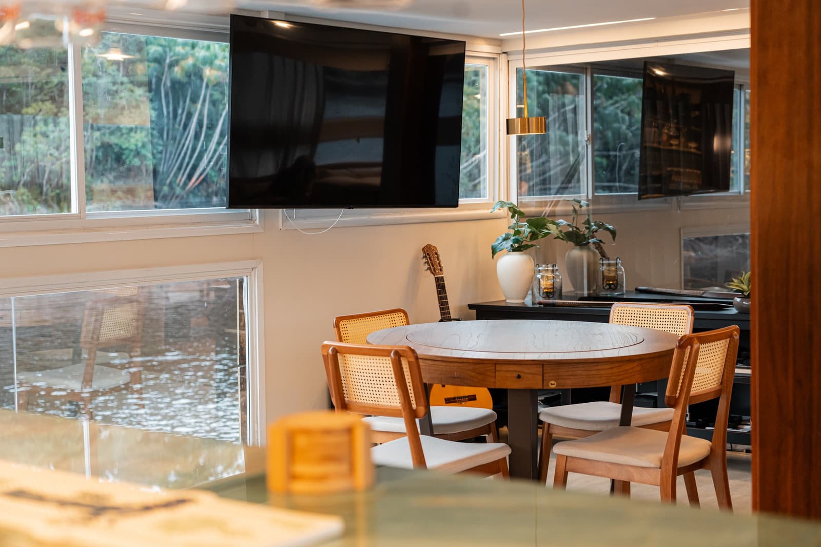 A round table, a guitar in the corner, and the river just outside the window. Round dining table with cane-back chairs aboard the untamed amazon cruise vessel, with a mounted flat-screen TV and lush green forest visible through panoramic windows