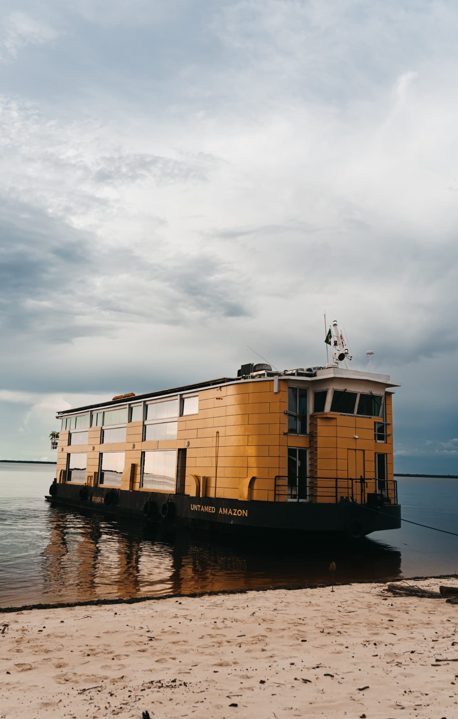 Das schwimmende Basislager, bereit am Ufer des Amazonas. Das goldgelbe Flusskreuzfahrtschiff Untamed Amazon liegt an einem Sandstrand am Amazonas unter einem dramatisch bewölkten Himmel