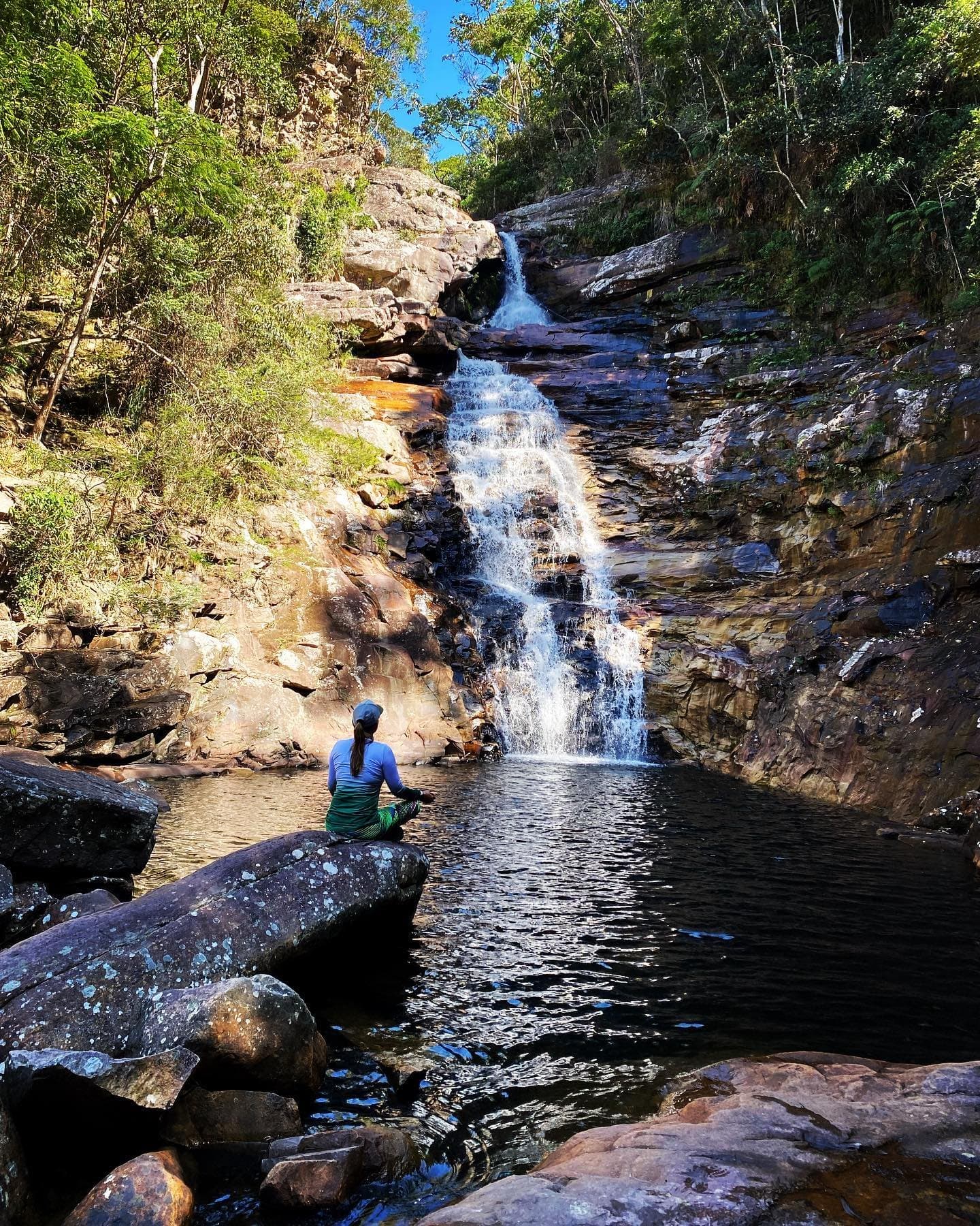 Às vezes a trilha termina aqui, numa pedra e num silêncio de água. Uma caminhante sentada em uma rocha à beira da piscina natural de uma cachoeira em degraus que desce por paredões de arenito no Trek Vale do Pati, cercada por mata atlântica sob céu azul