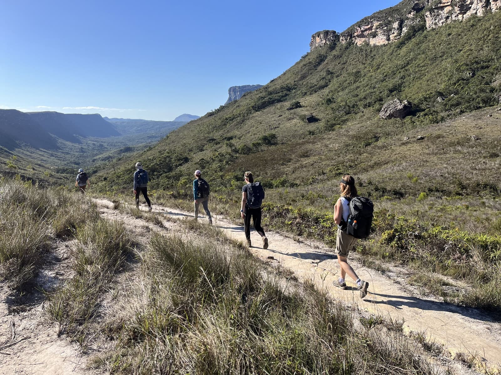 O vale do Pati não se revela de uma vez, ele vai te contando aos poucos. Cinco caminhantes com mochilas percorrem em fila um trilho de pedra arenosa no Vale do Pati, com vistas panorâmicas do vale e escarpas de arenito sob um céu azul sem nuvens