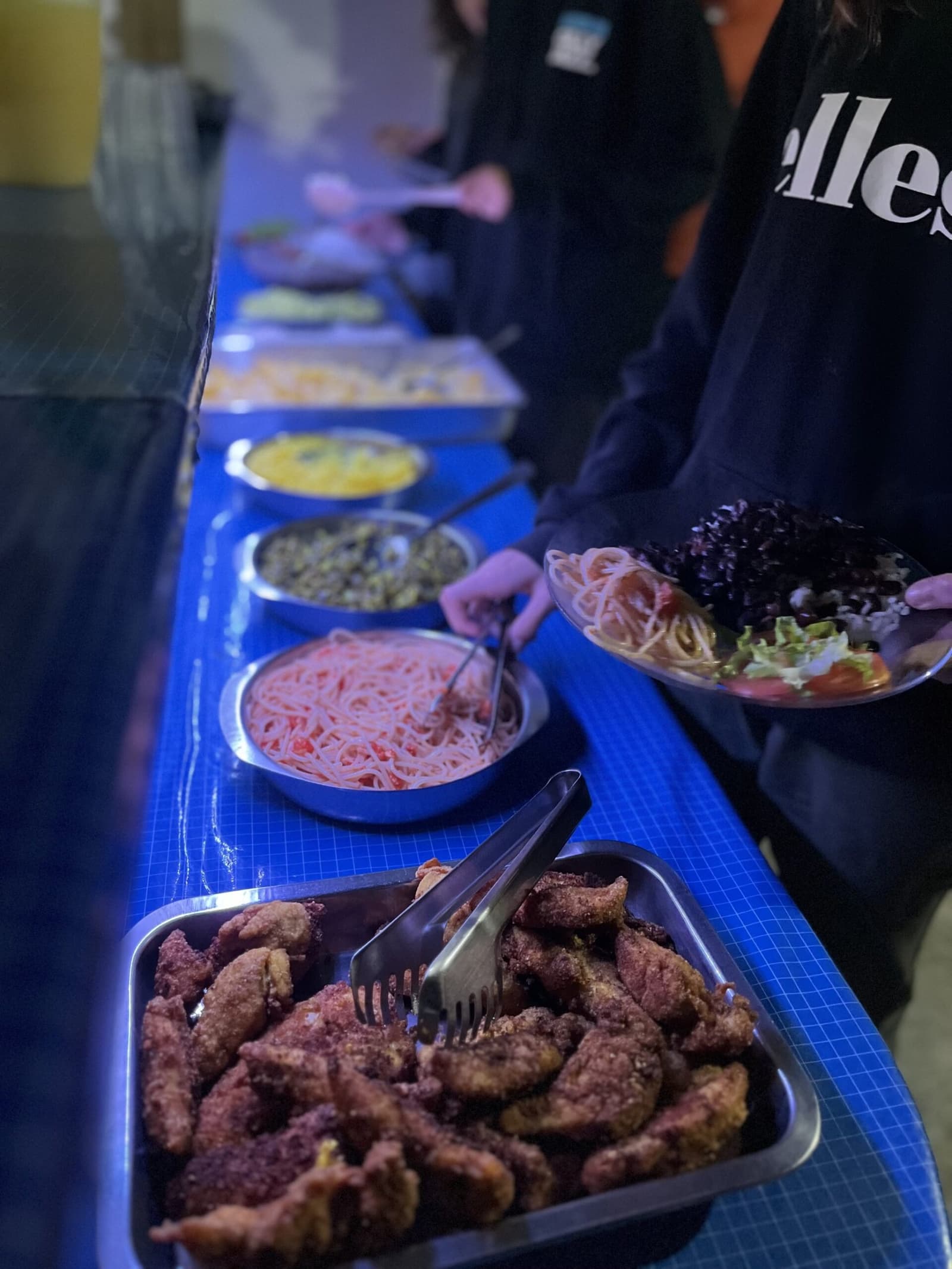 Comida simples, trilha longa, prato cheio. Uma longa mesa de buffet com pratos caseiros — carne frita empanada, macarrão ao molho de tomate, lentilhas e salada fresca — servidos em mesa com toalha azul no jantar de uma família anfitriã no Vale do Pati