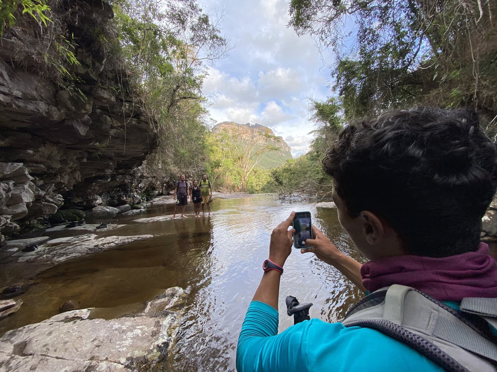 Água no joelho, memória garantida, assim se faz uma trilha no Vale do Pati. Um caminhante fotografa dois trilheiros sorrindo dentro de um rio raso em um canyon rochoso no Vale do Pati Trek, com paredões de arenito e uma chapada ao fundo