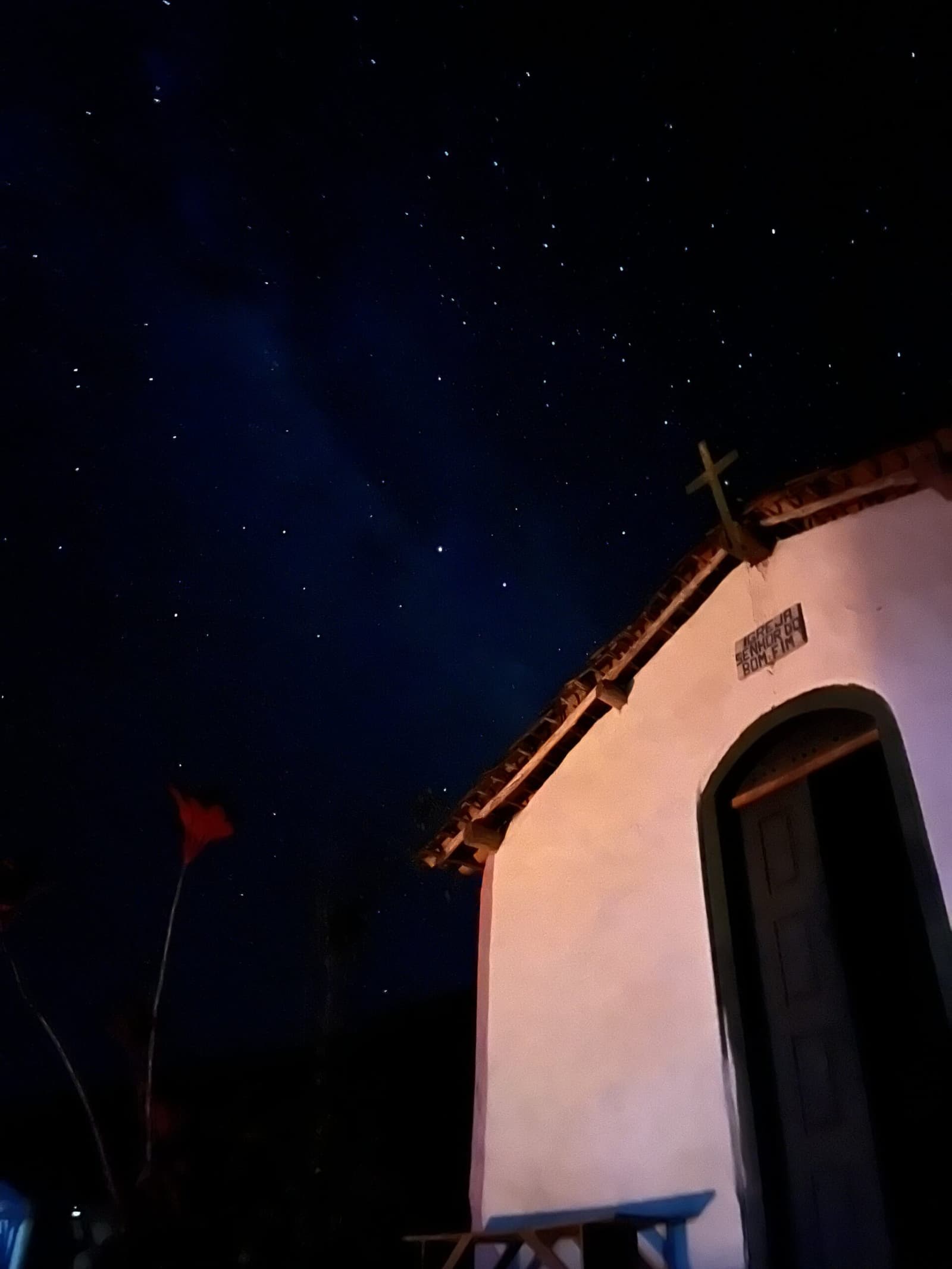 Paredes antigas, estrelas mais antigas ainda. Tem noites na Chapada que a gente para e fica. A Igreja Senhor do Bonfim, pequena e caiada, vista de baixo ângulo sob um céu repleto de estrelas durante o Trek Vale do Pati, com uma cruz de madeira recortada no azul profundo da noite