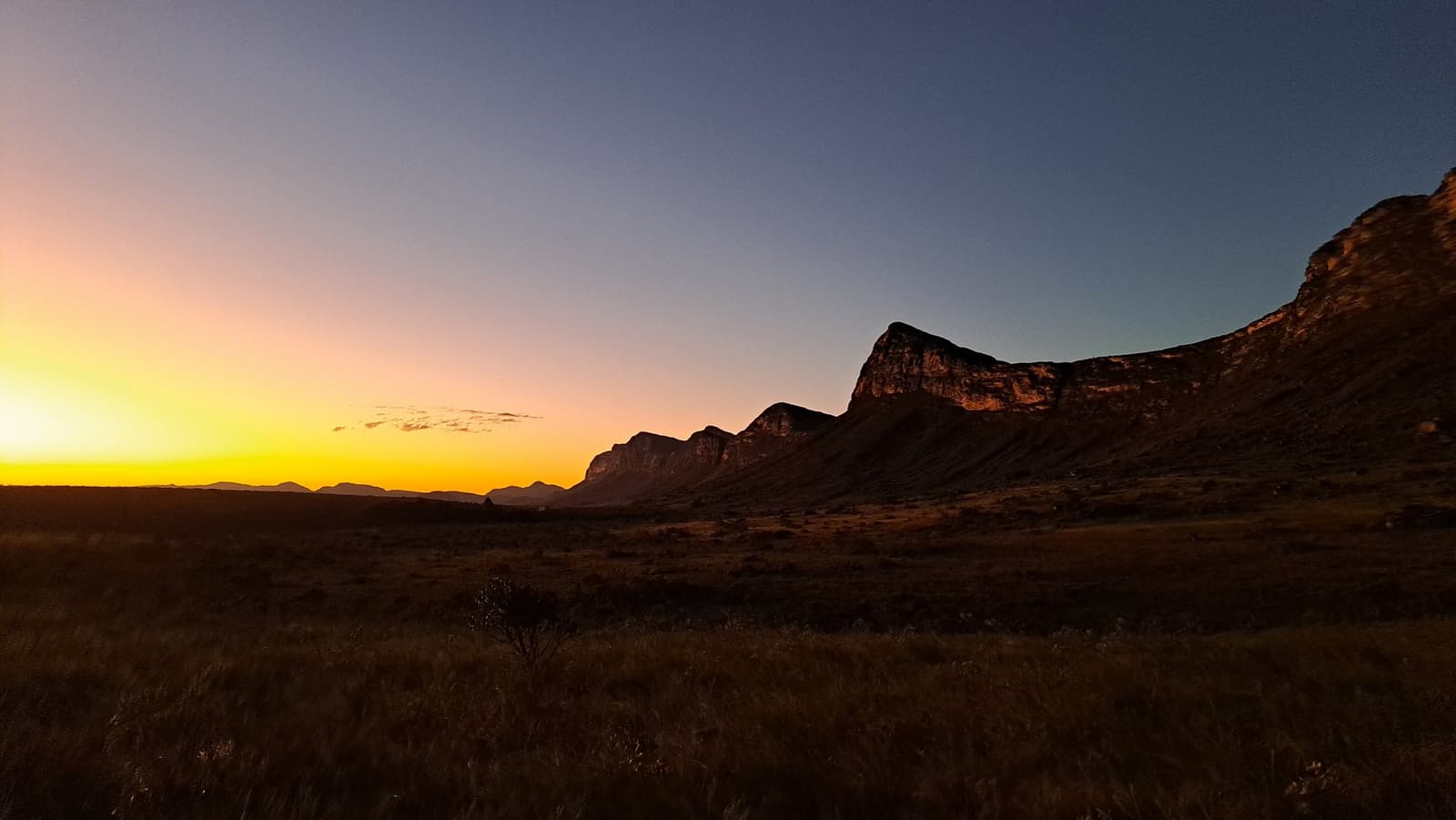 A Chapada despede o dia do jeito dela: sem pressa, com tudo. Pôr do sol em tons laranja e dourado sobre uma vasta planície aberta com uma escarpa rochosa em silhueta contra o céu azul profundo no trek Vale do Pati