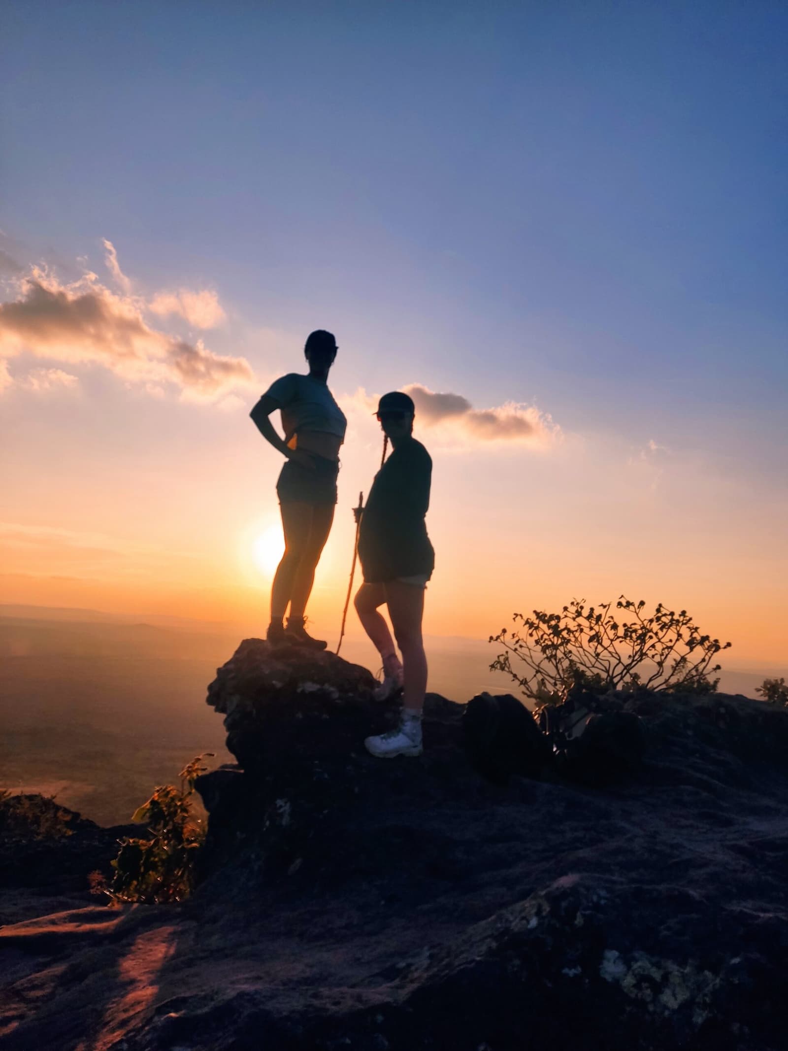 Chegou lá em cima e o sertão respondeu. Duas caminhantes em silhueta sobre uma rocha no topo de uma trilha do Vale do Pati, com o pôr do sol alaranjado iluminando um vasto panorama de planícies