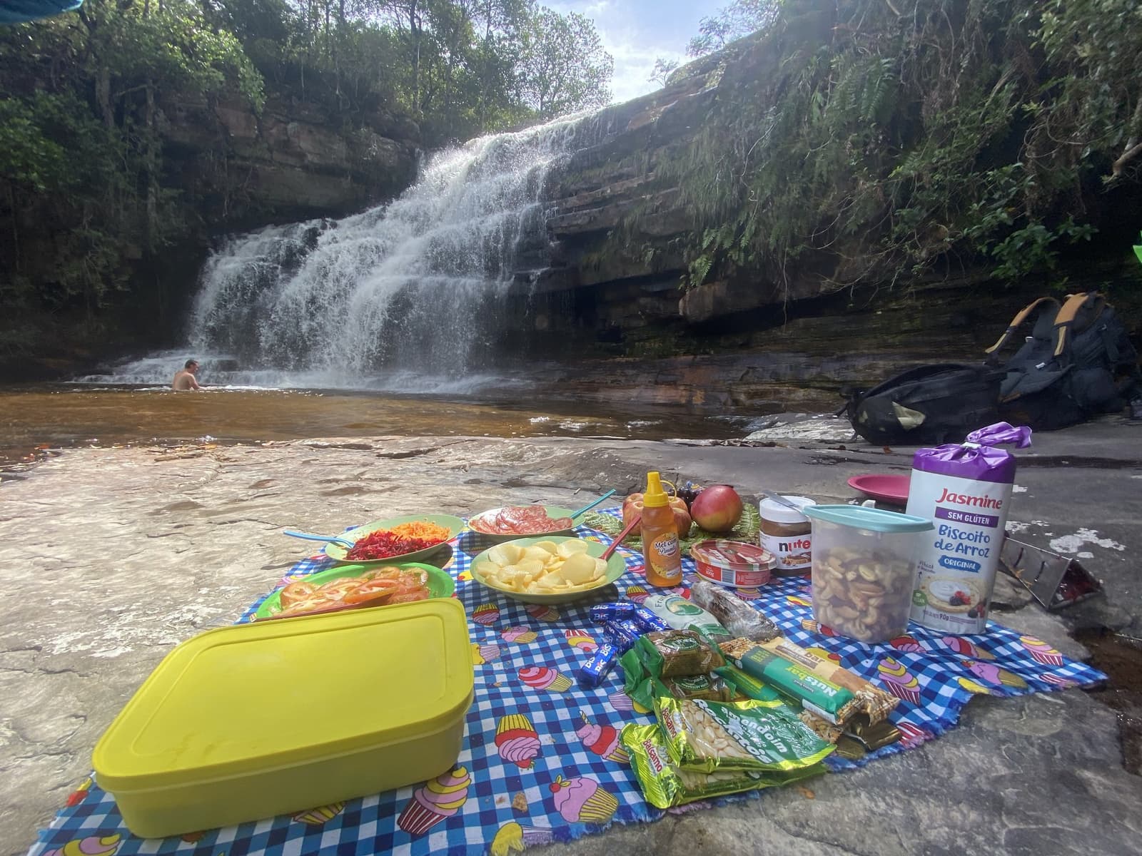 Almoço simples, cenário que nenhum restaurante entrega. Piquenique colorido em toalha xadrez sobre rochas planas ao lado de uma cachoeira na trilha do Vale do Pati, com saladas, frutas, mel e lanches, enquanto um banhista aproveita a piscina natural ao fundo