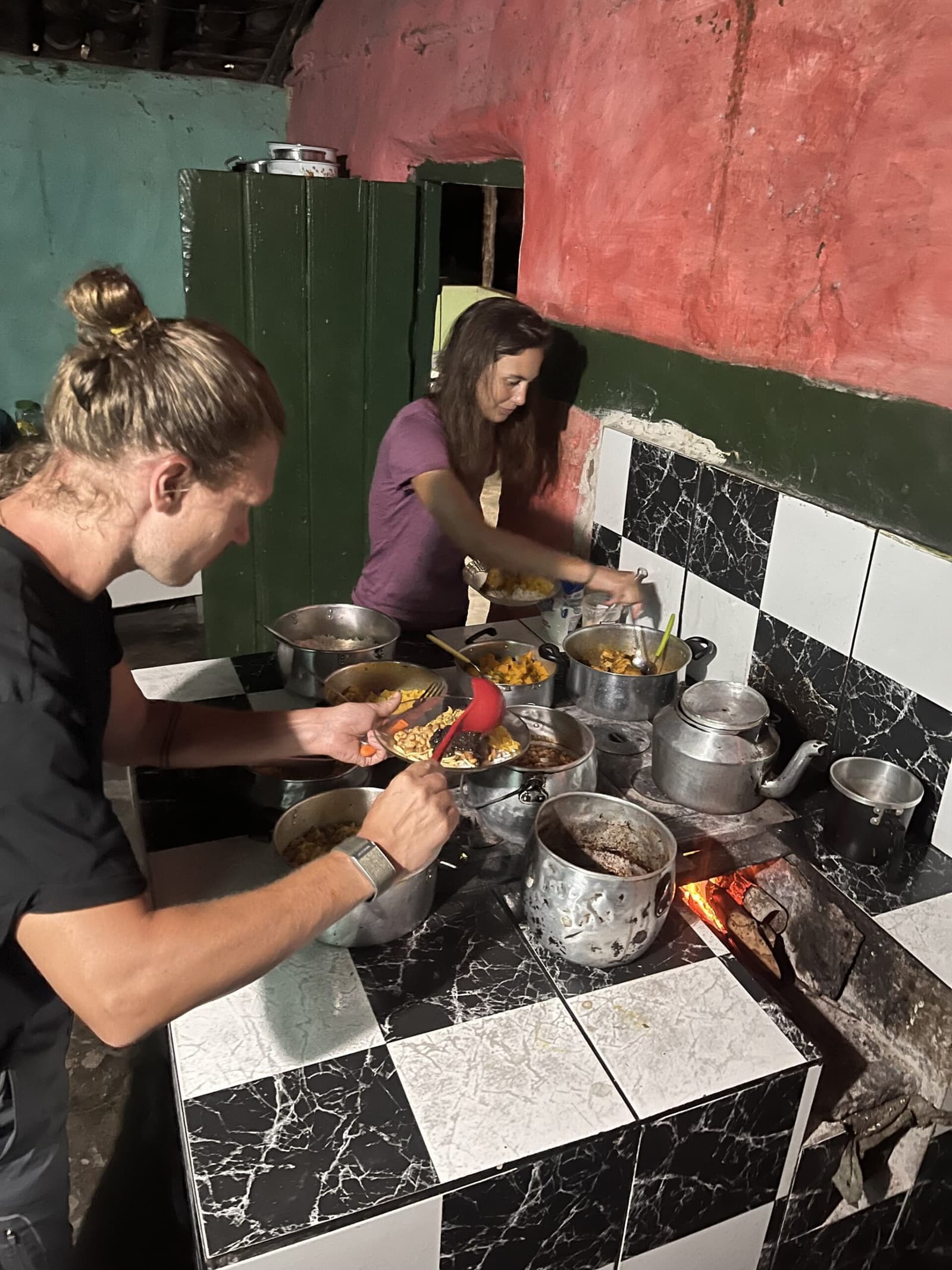 Fogão a lenha, panelas cheias e o cansaço gostoso de quem andou muito. Dois caminhantes se servindo de várias panelas fervendo num fogão a lenha em uma cozinha rústica durante o jantar em família na Vale do Pati Trek