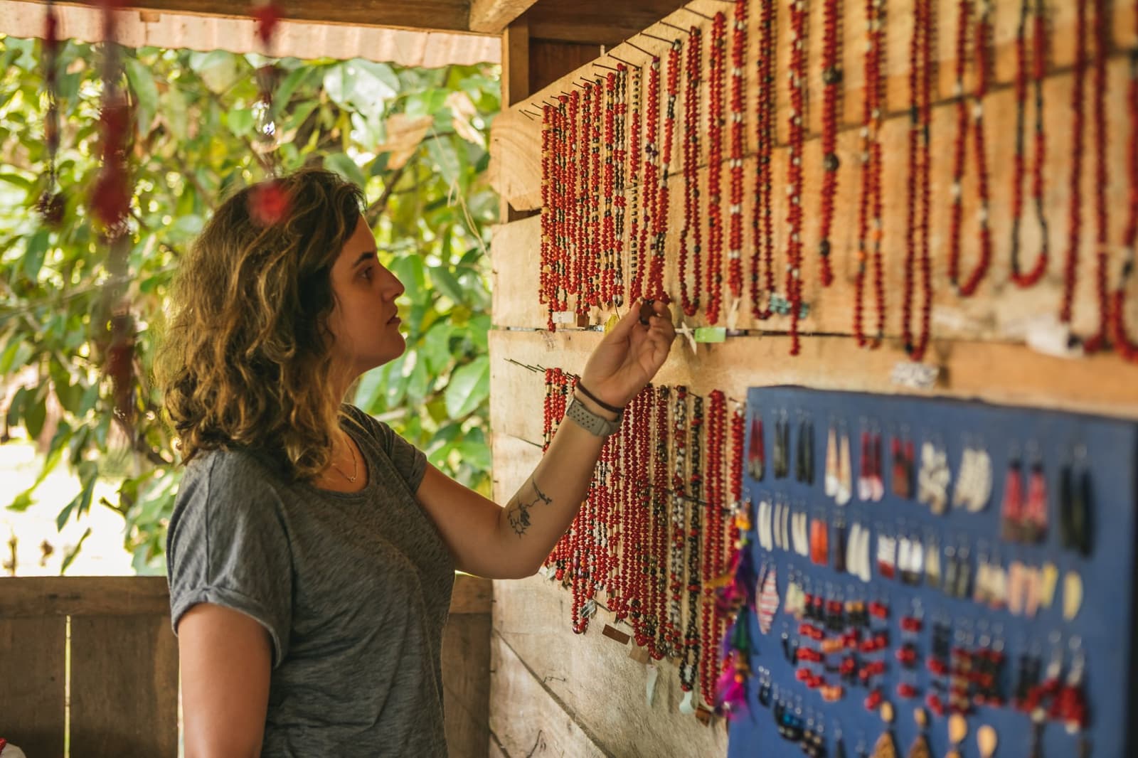 Local seed beads: the kind of souvenir with a story attached. Photo: Guilherme Gomes A woman browses rows of red seed bead necklaces and small earrings displayed on a wooden wall at a riverside community craft stall during an amazon rainforest ecolodge trip in brazil
