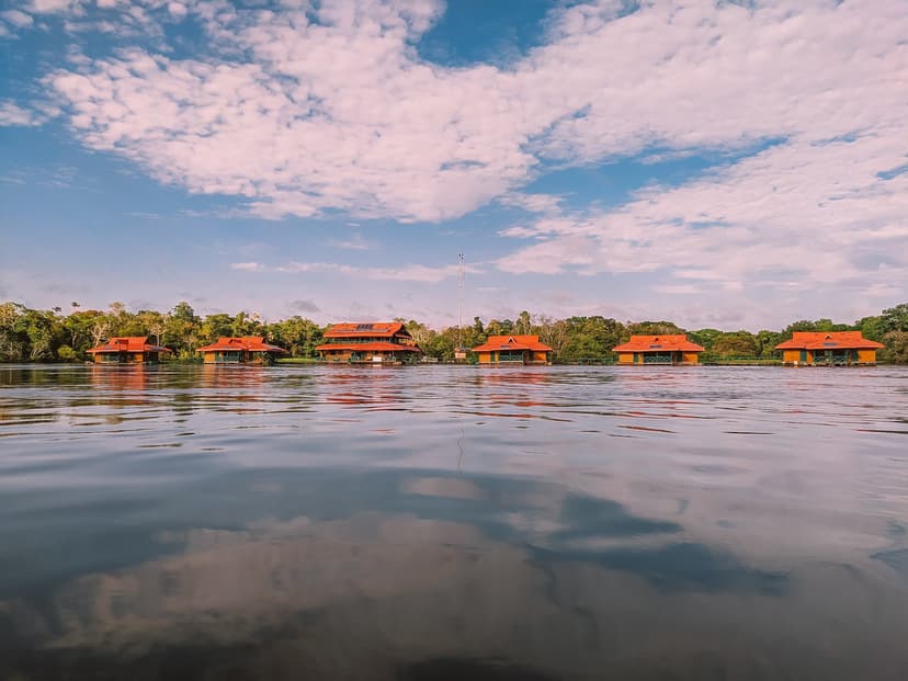 A row of red-roofed wooden bungalows at the Uakari Lodge, an amazon rainforest ecolodge trip in Brazil, seen from water level with clouds reflected in the calm river and dense forest behind A row of red-roofed wooden bungalows at the Uakari Lodge, an amazon rainforest ecolodge trip in Brazil, seen from water level with clouds reflected in the calm river and dense forest behind