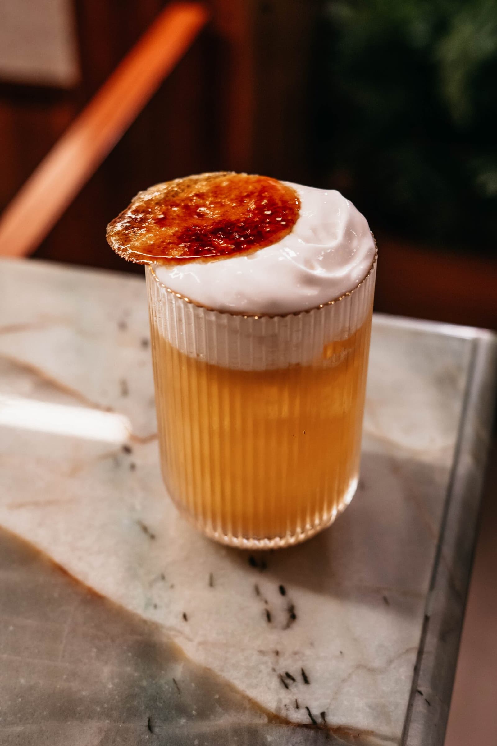 Wenn die Flamme das letzte Wort hat. Ein gerilltes Glas mit goldenem Cocktail, dichtem weißem Schaum und einem flambiertem Karamellscheiben-Garnish auf einer Marmoroberfläche an Bord einer Luxus-Amazonas-Kreuzfahrt in Brasilien