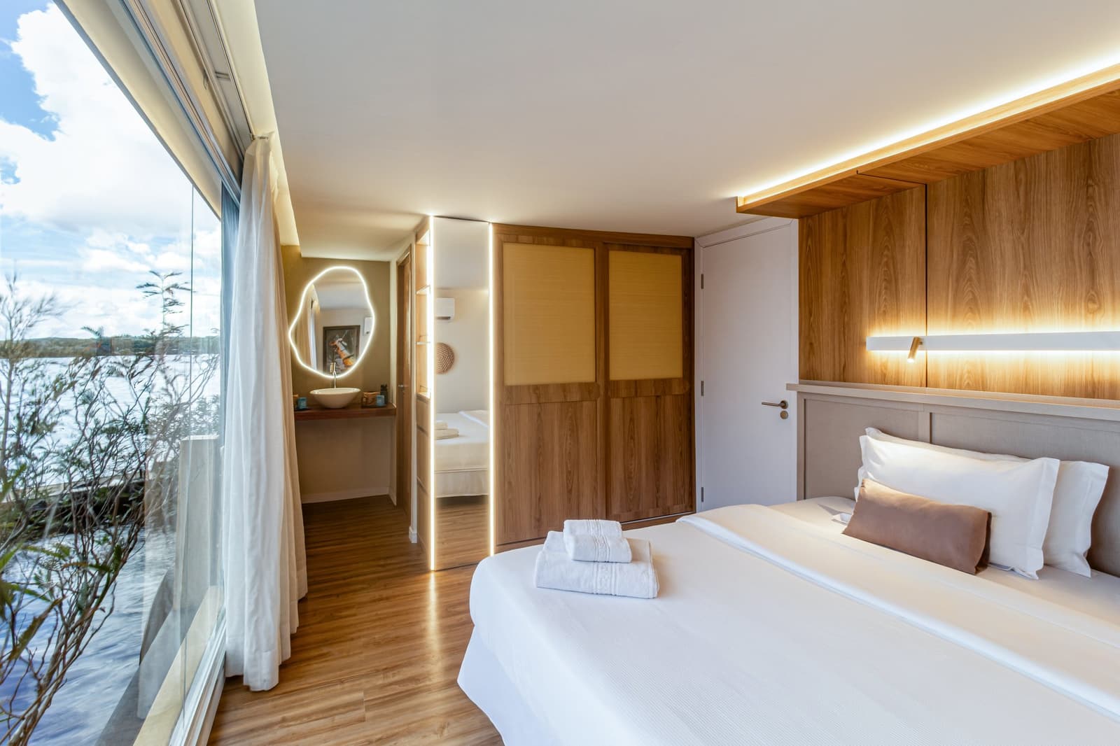Holz, weiches Licht und der Fluss direkt vor dem Fenster. Photo: STUDIO CREATE Helle Suite auf einer Luxus-Amazonas-Kreuzfahrt in Brasilien mit Doppelbett und weißer Bettwäsche, warmem Holzkopfteil mit LED-Beleuchtung, raumhohen Fenstern mit Blick auf einen breiten Fluss und einem offenen Bereich mit beleuchtetem Schminkspiegel