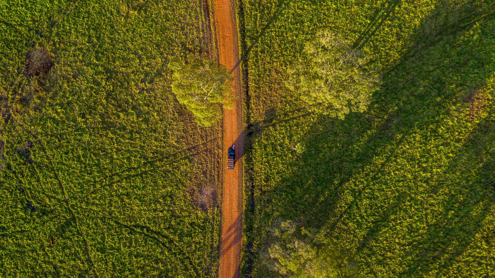 Fifty-three thousand hectares, one red road, and you somewhere in the middle of it. Aerial top-down view of a vehicle on a red dirt track cutting through dense green scrubland on a luxury Pantanal and Amazon trip