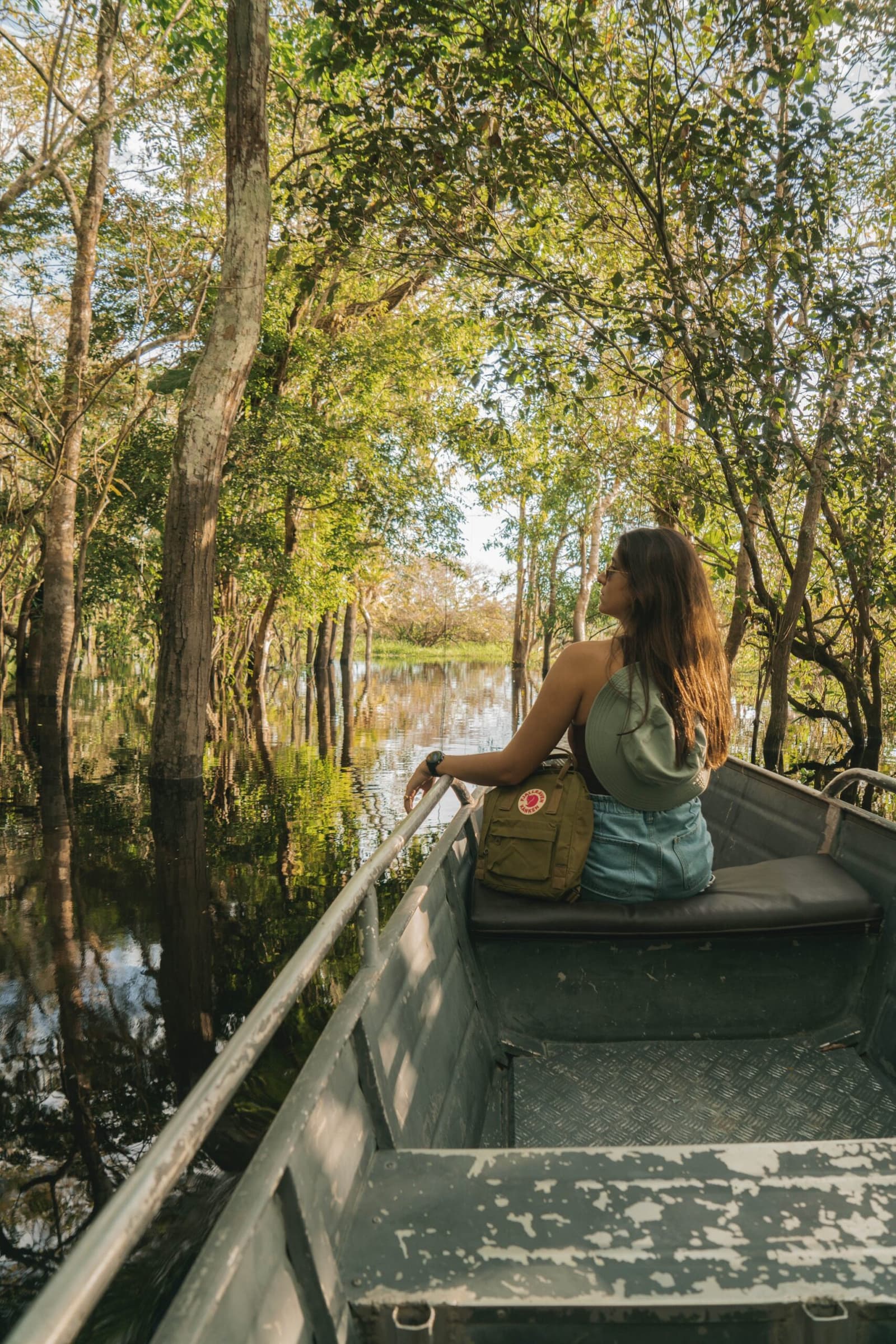Quiet water, tall trees, and nowhere else you need to be. Photo: Yuri Panichi A woman sits at the bow of a metal boat on an amazon and pantanal tour, looking out at a calm flooded forest channel lined with tall trees reflected in still water