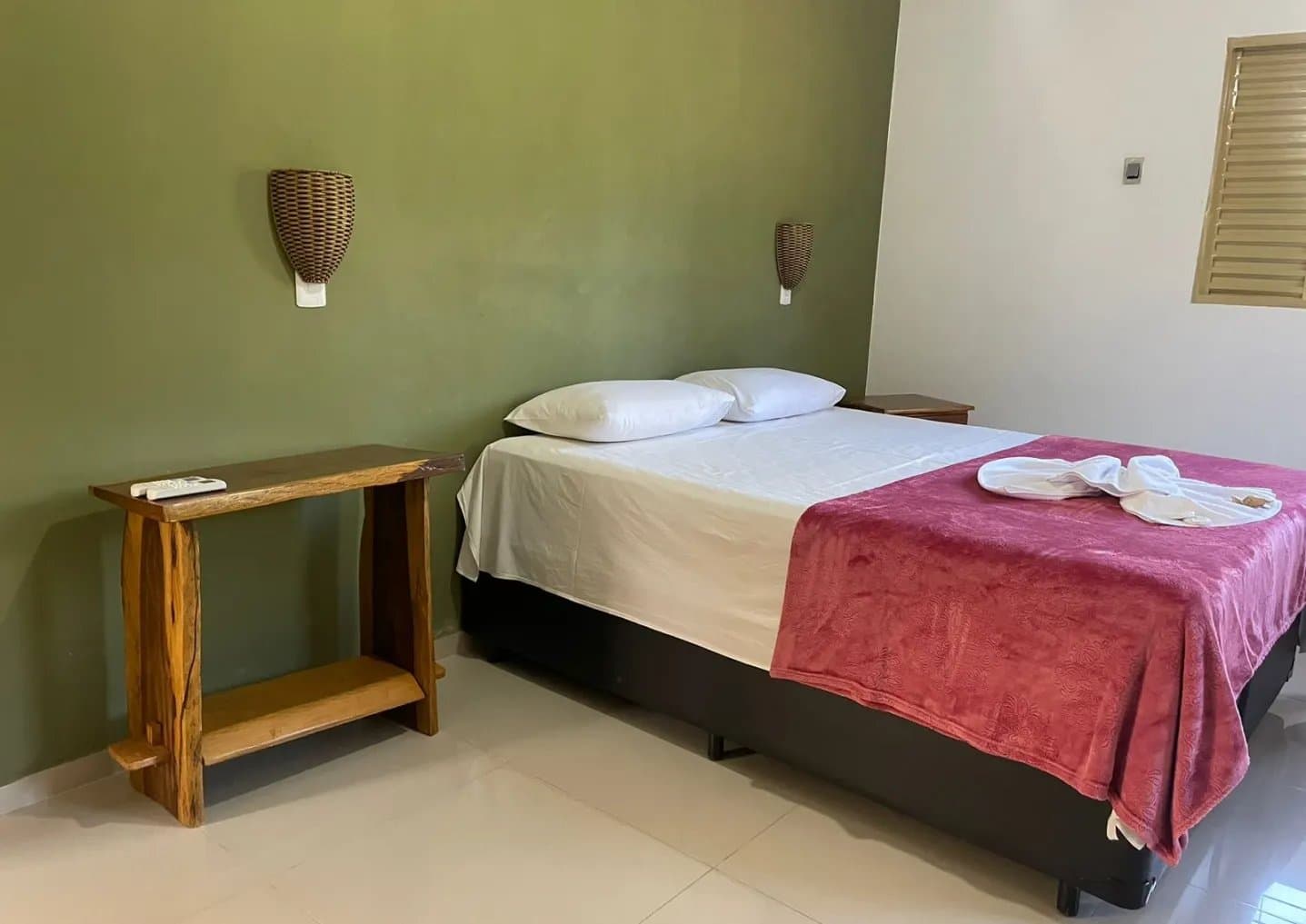 Earthy green walls, folded towels, and a good night ahead of you. Simply furnished double room on an amazon and pantanal tour, featuring a double bed with white linens and a red throw, wicker wall sconces, and a handcrafted wooden side table
