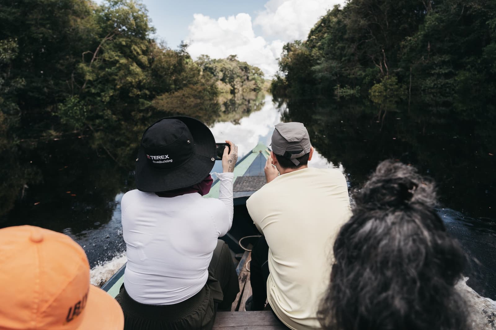 Schmal am Anfang, dann öffnet sich alles, der Kanal und der Blick. Vier Reisende sitzen auf einem Holzboot auf einem Luxus-Amazonas-Kreuzfahrtschiff in Brasilien und fahren durch einen schmalen Schwarzwasserkanal, der von dichtem Regenwald gesäumt ist; eine Person fotografiert mit dem Smartphone voraus