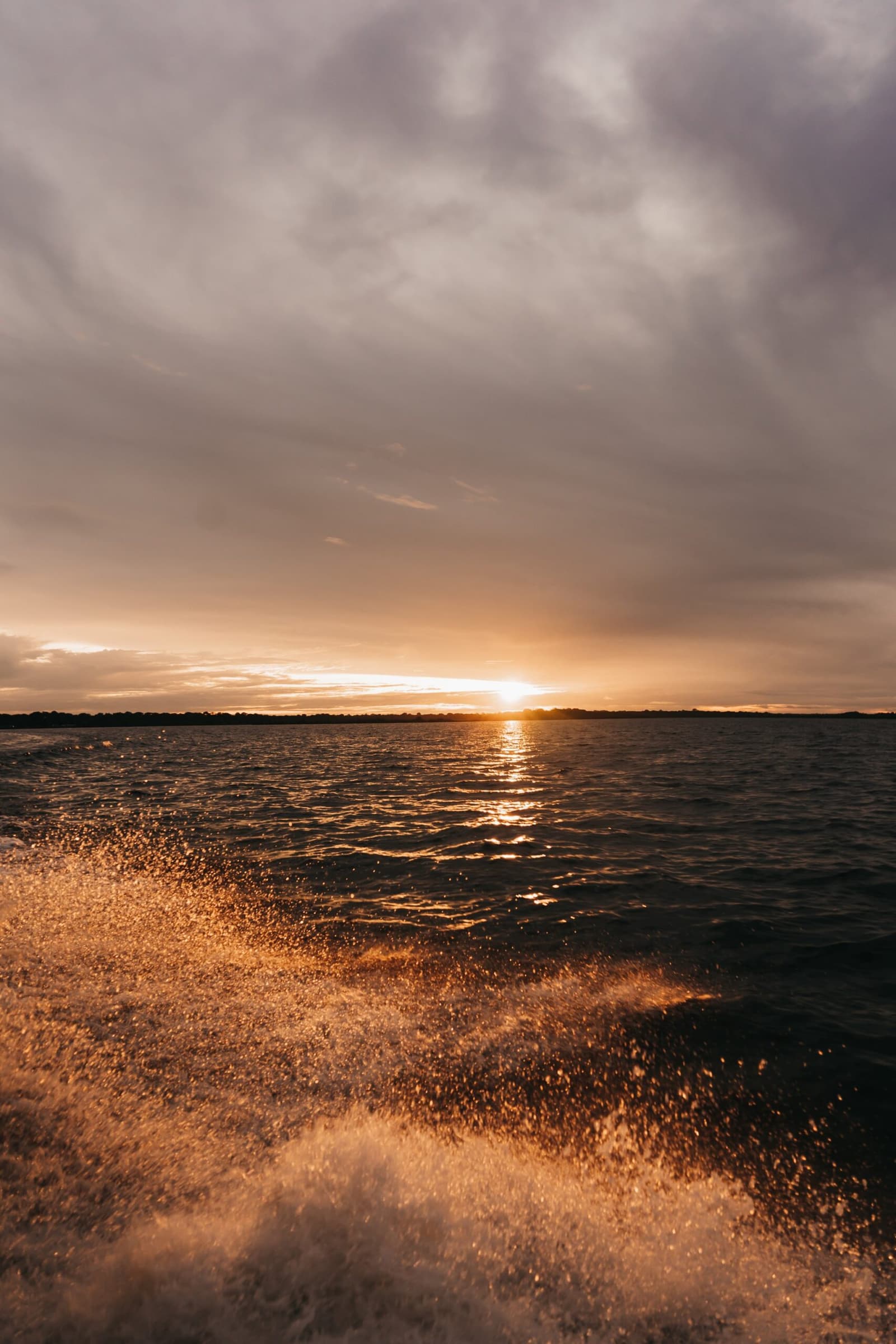 Wenn das Licht so fällt, legt man die Kamera erst gar nicht weg. Goldenes Bugwasser eines Bootes vor einem Sonnenuntergang über einem breiten Fluss, mit einer fernen Baumsilhouette unter bewölktem Himmel auf einer Amazonas-Kreuzfahrt in Brasilien