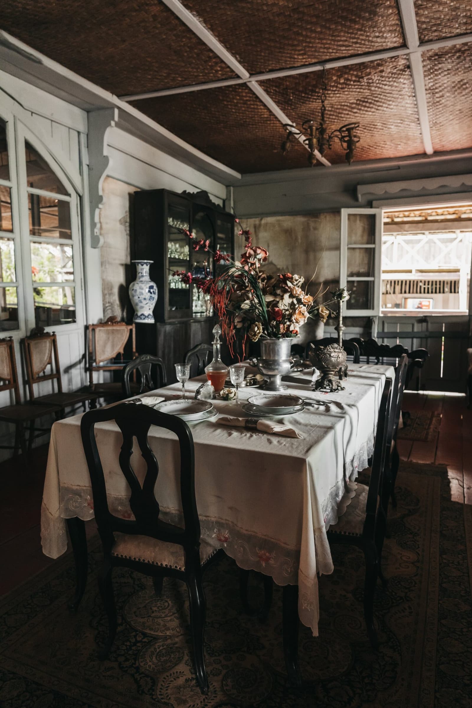 Ein Tisch, an dem die Gummibaum-Ära noch zwischen den Wänden hängt. Historischer Speisesaal mit gedecktem Tisch, weißem Leinentischtuch, Silber- und Kristallgeschirr, dunklen Holzstühlen, blau-weißer Porzellanvase und gewebter Decke in weichem Tageslicht