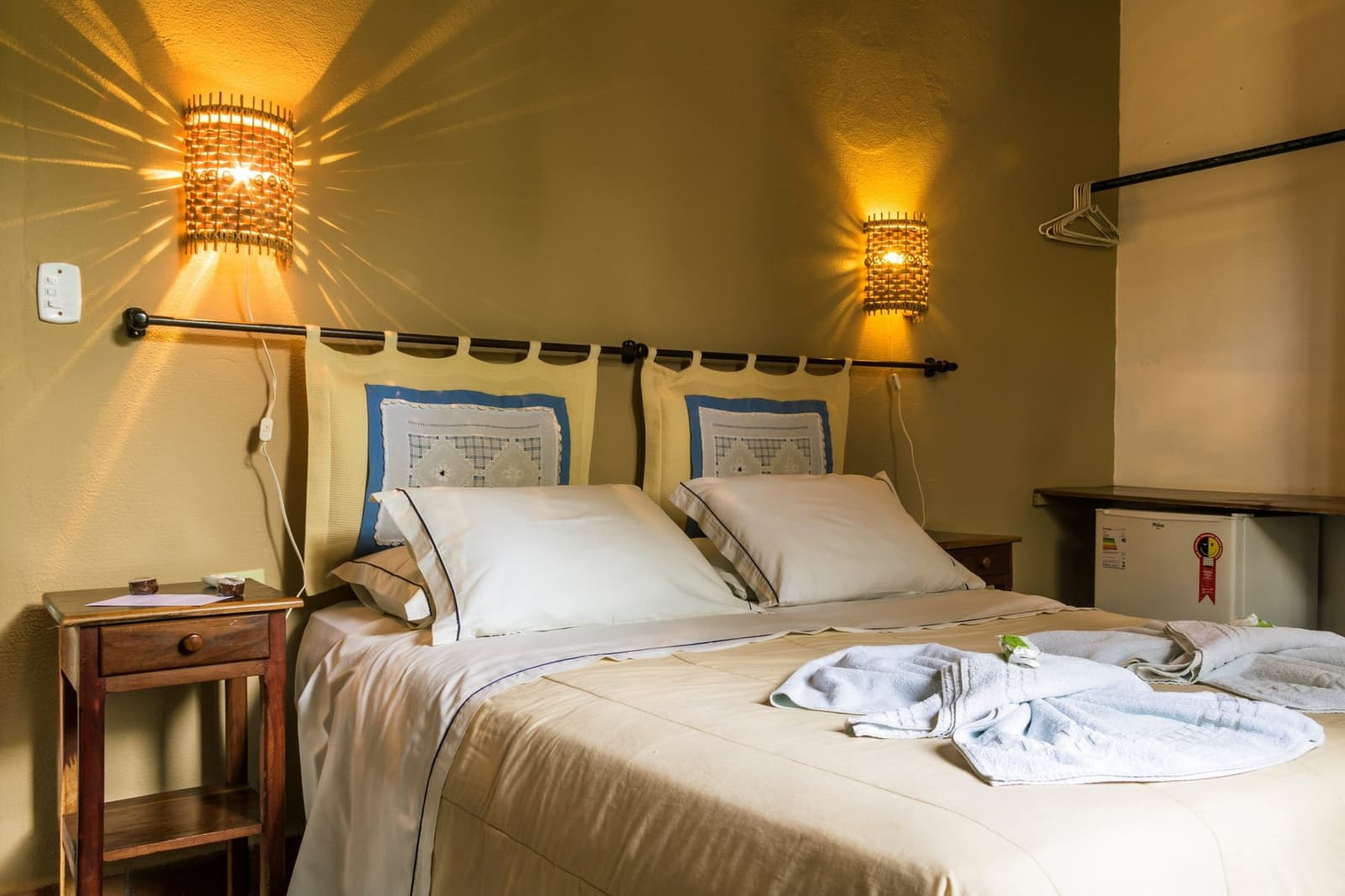After a night safari, this room earns its welcome. Cozy double room at a Pantanal lodge on the pantanal bonito tour, featuring warm woven-bamboo wall sconces, cream bedding with blue accent pillowcases, folded bath robes, and a wooden bedside table