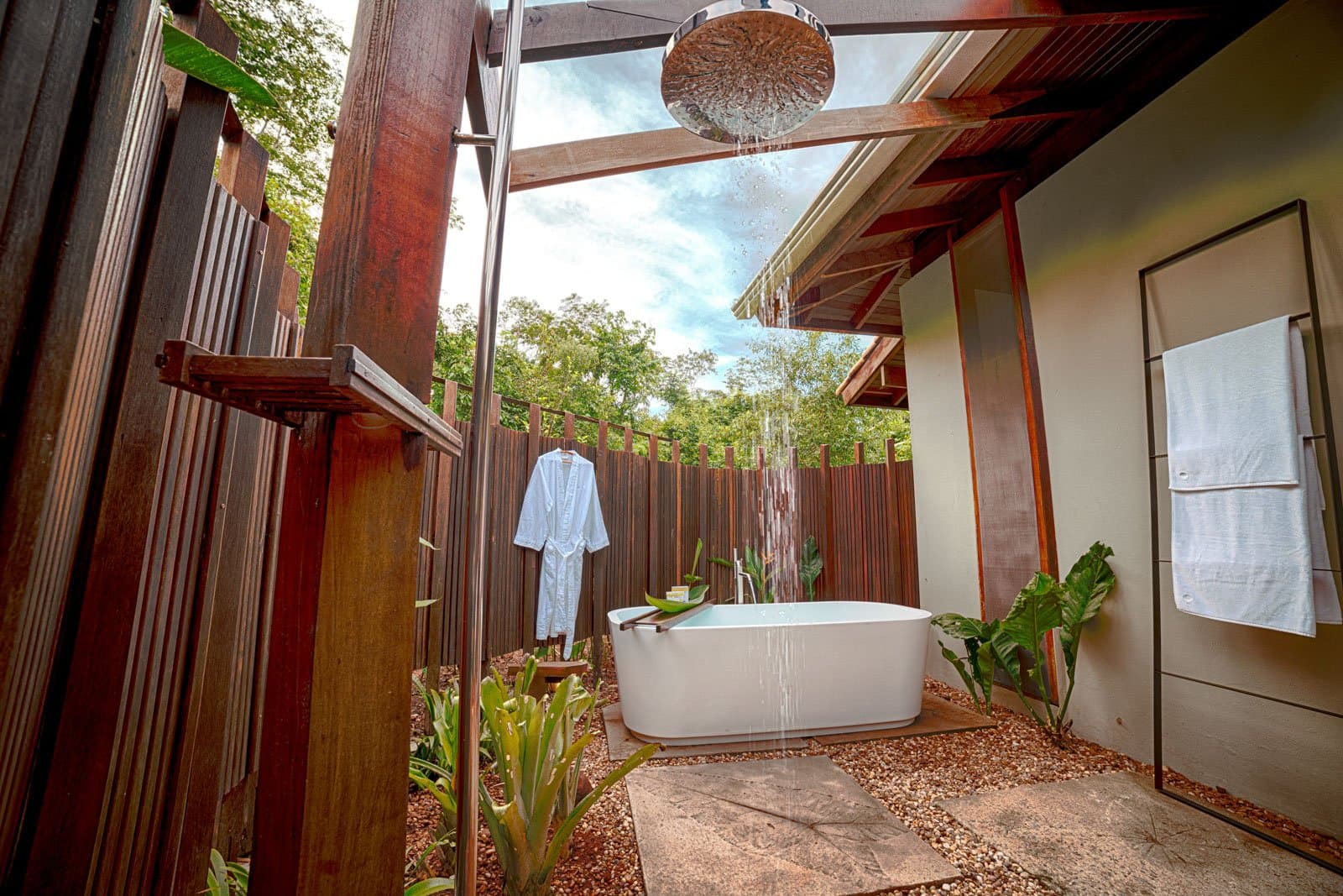 Rain shower, treetops, and a bathtub with no ceiling to speak of. Photo: Samuel Melim Open-air bathroom at a luxury Pantanal and Amazon trip lodge, featuring a freestanding white soaking tub under a rain showerhead, framed by dark hardwood screens, tropical plants, and a sky-open roof