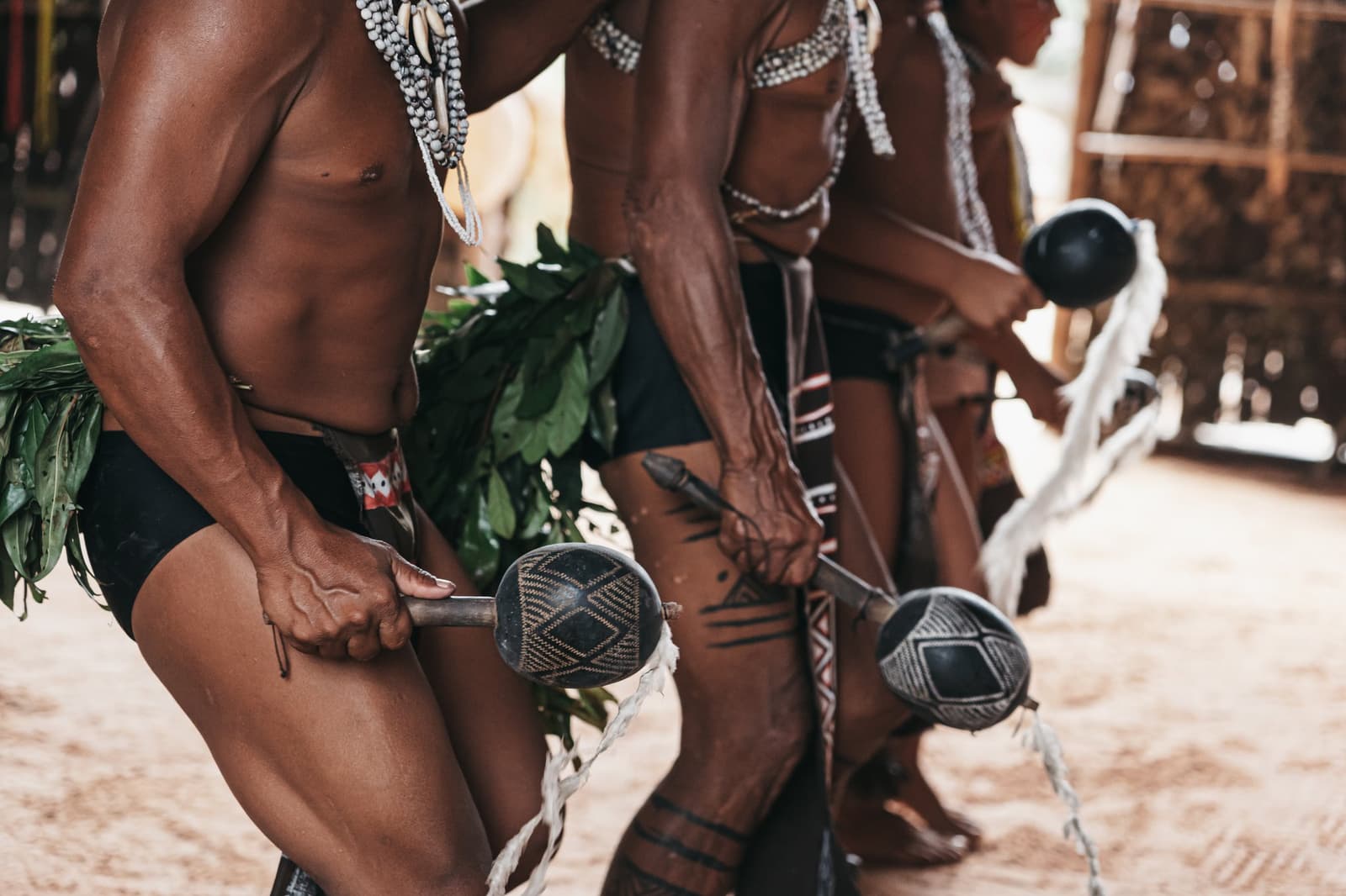 Rasseln, Tätowierungen und ein Rhythmus, der keine Übersetzung braucht. Nahaufnahme indigener Männer, die bei einer traditionellen Zeremonie verzierte Kürbisrasseln halten, mit Perlenketten, Blattröcken und Körpertätowierungen