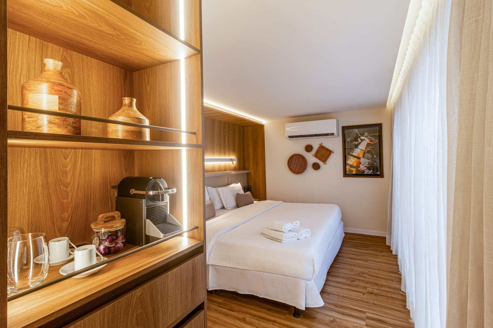 Ein Kaffee wartet schon, bevor der Tag überhaupt begonnen hat. Photo: STUDIO CREATE Eine komfortabel eingerichtete Kabine auf einem Luxus-Amazonas-Kreuzfahrtschiff in Brasilien mit einem Doppelbett in weißer Bettwäsche, gefalteten Handtüchern, einer beleuchteten Holzregalwand mit Kapselkaffeemaschine, Keramiktassen und dekorativen Tonvasen sowie gewebten Wandkörben und hellen Vorhängen