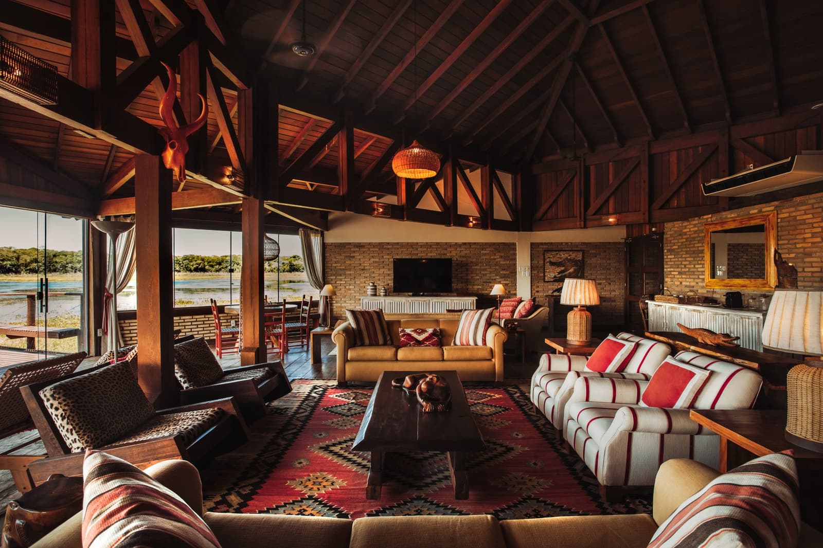 A room that earns its place, the river is right there, through those doors. Photo: Felipe Castellari Spacious lodge common room on a luxury Pantanal and Amazon trip, featuring exposed dark-wood roof trusses, a woven pendant lamp, kilim rug, leather and striped sofas, animal-skull wall décor, and open glass doors framing a river view beyond the deck