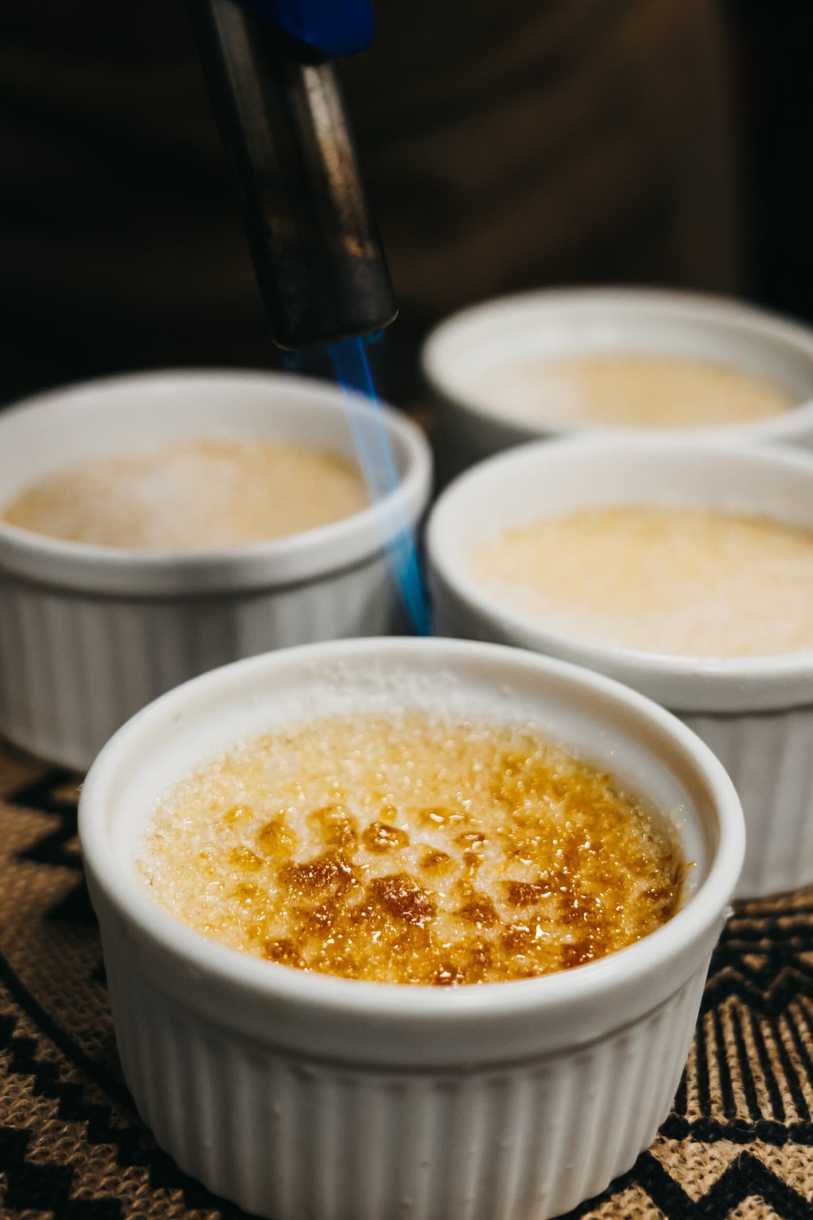 Crème brûlée, mitten im Regenwald. Das Knacken der Kruste macht das Erlebnis vollständig. Eine Küchenbrennerflamme karamellisiert die Zuckerkruste einer Crème brûlée in einem weißen Förmchen; drei weitere unfertige Förmchen warten im Hintergrund auf einem gemusterten Textil auf einer Amazonas-Kreuzfahrt in Brasilien.
