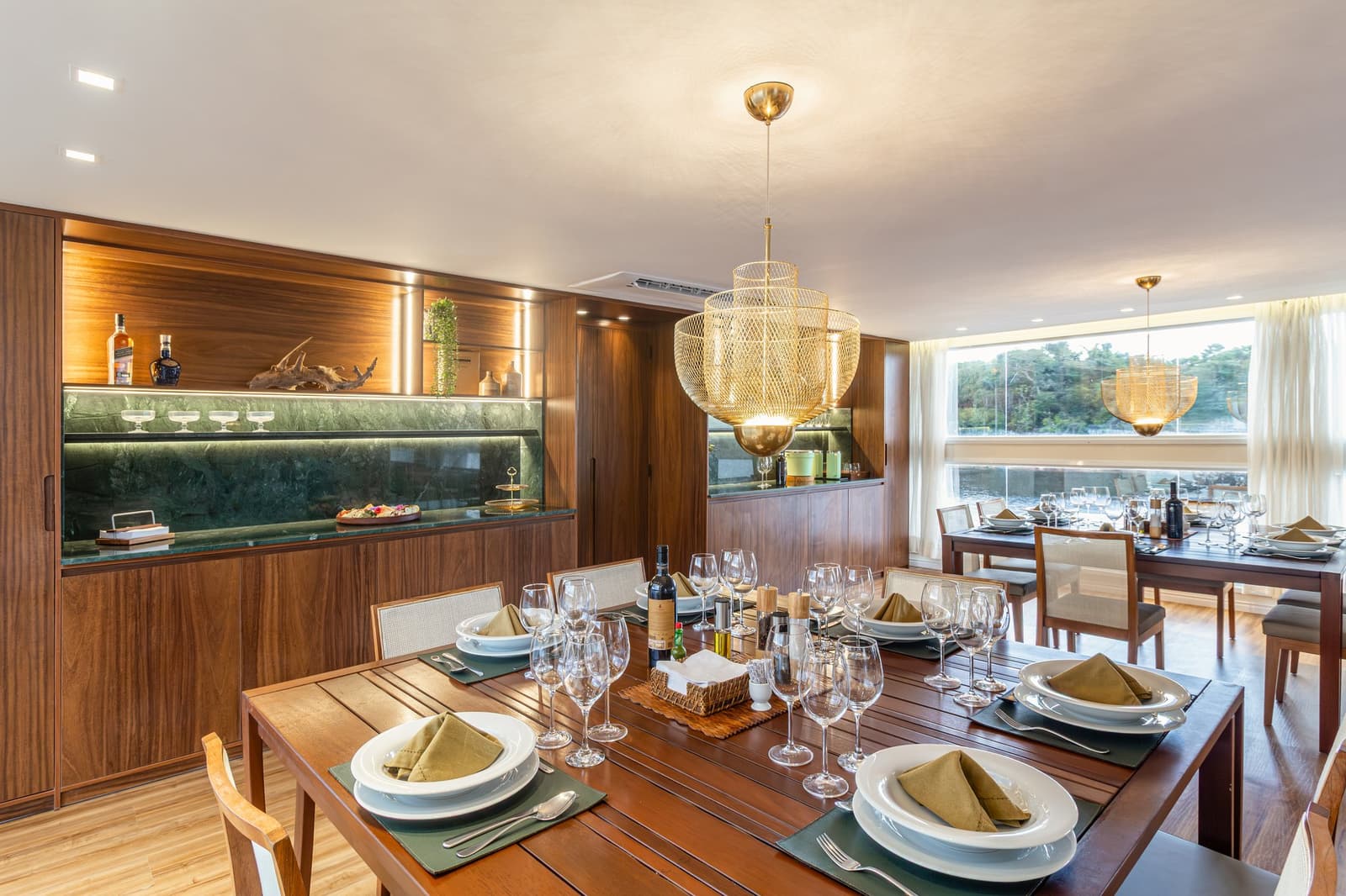 Wenn der Amazonas am Fenster vorbeizieht, schmeckt das Essen gleich besser. Photo: STUDIO CREATE Elegant gedeckter Speisesaal an Bord einer Luxus-Amazonas-Kreuzfahrt in Brasilien mit zwei Holztischen, weißem Geschirr, olivfarbenen Leinenservietten, mehreren Weingläsern, einer beleuchteten Walnuss-Bar mit grüner Marmorplatte und großen Fenstern zum Fluss