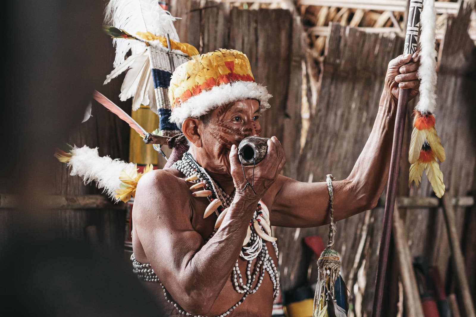 Jahrzehnte gelebte Tradition, konzentriert in einem einzigen Moment. Ein indigener Ältester mit Federkopfschmuck und Knochenketten bläst während einer Gemeinschaftszeremonie in ein kleines Kalebassen-Instrument und hält dabei einen verzierten Zeremonialstab in einer strohgedeckten Hütte.