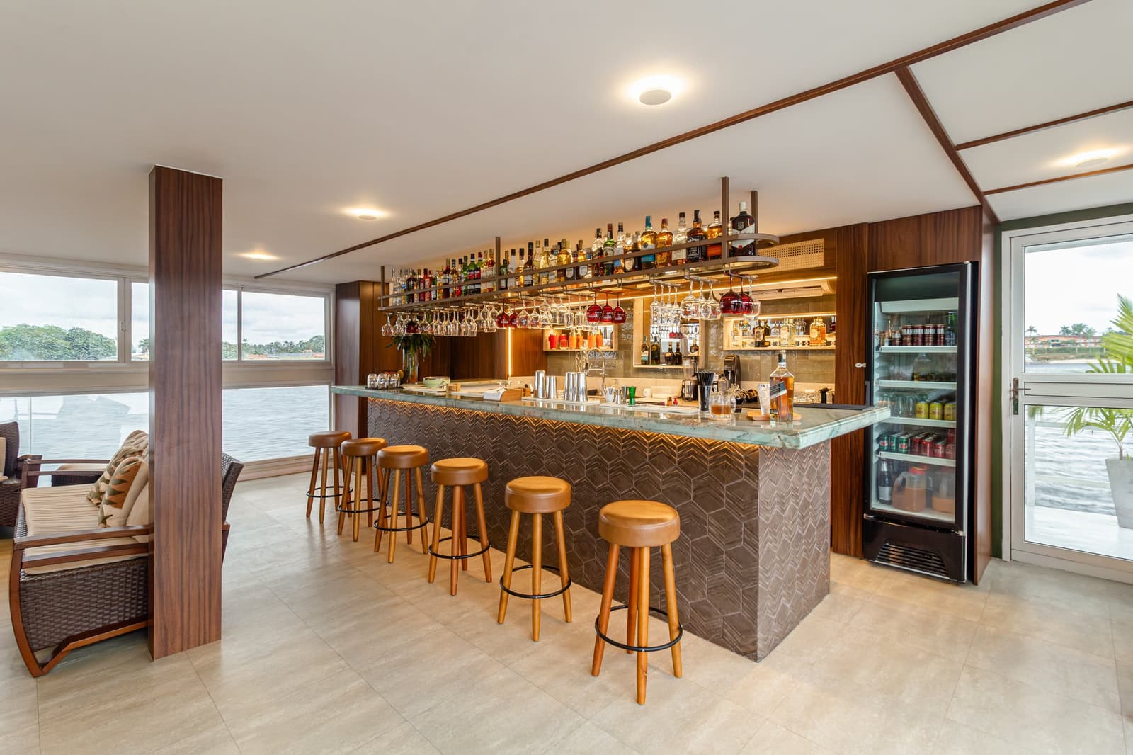 Die Bar ist bereit, der Fluss fließt draußen vorbei, kein schlechter Platz für den Feierabend. Photo: STUDIO CREATE Gut bestückter Bar-Bereich auf einer Luxus-Amazonas-Kreuzfahrt in Brasilien, mit grüner Marmortheke, beleuchteten Spirituosenregalen, hängenden Weingläsern, Lederhockern und Panoramafenstern mit Blick auf den Fluss
