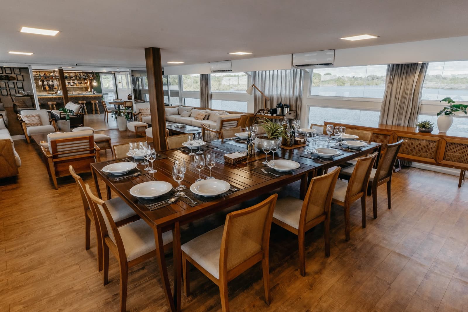 Gedeckter Tisch, draußen der Rio Negro. So sieht Mittagessen hier aus. Formal gedeckter Esstisch mit weißem Porzellan und Weingläsern auf einer Luxus-Amazonas-Kreuzfahrt in Brasilien, dahinter ein Loungebereich und große Fenster mit Blick auf den Fluss