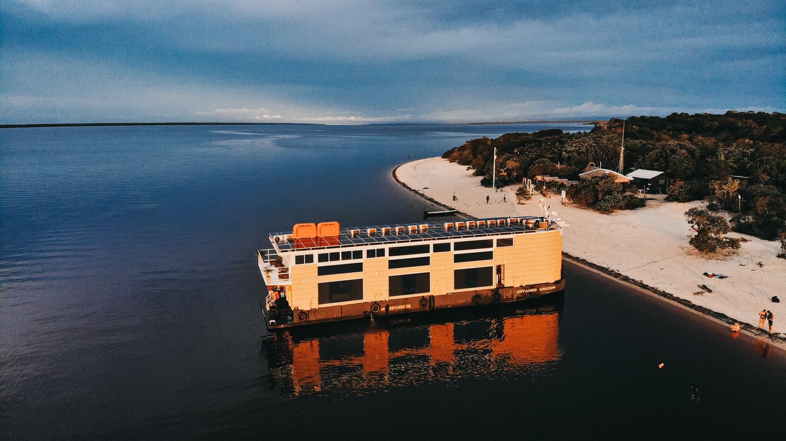 Vier Nächte an Bord, und der Strand liegt direkt vor der Gangway. Luftaufnahme des solarbetriebenen Flussboots Untamed Amazon, das bei Sonnenuntergang neben einem weißen Flussstrand auf dem dunklen Rio Negro liegt, während einer Luxus-Amazonas-Kreuzfahrt in Brasilien