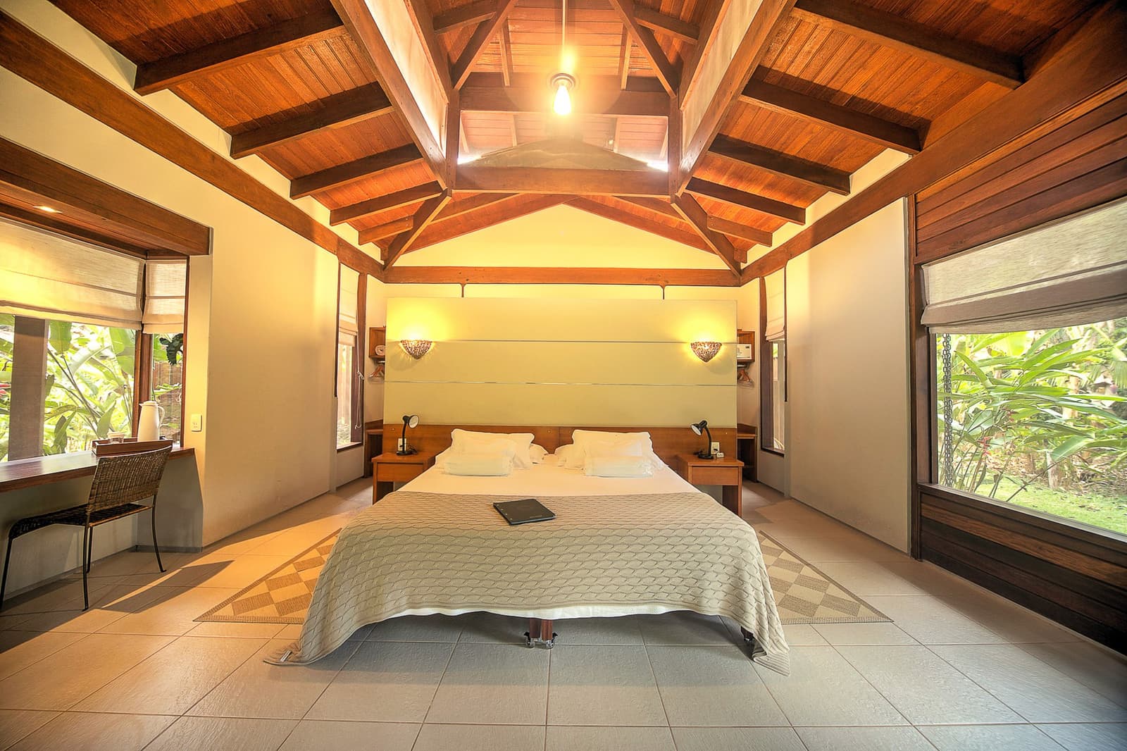 Tall ceilings, green just outside the glass, and the forest keeping you company all night. Photo: | SamuelMelim Photography Spacious lodge bedroom with a vaulted timber-beam ceiling, a double bed with white linens, warm wall sconces, and large windows framing lush tropical greenery — part of a luxury Pantanal and Amazon trip stay experience