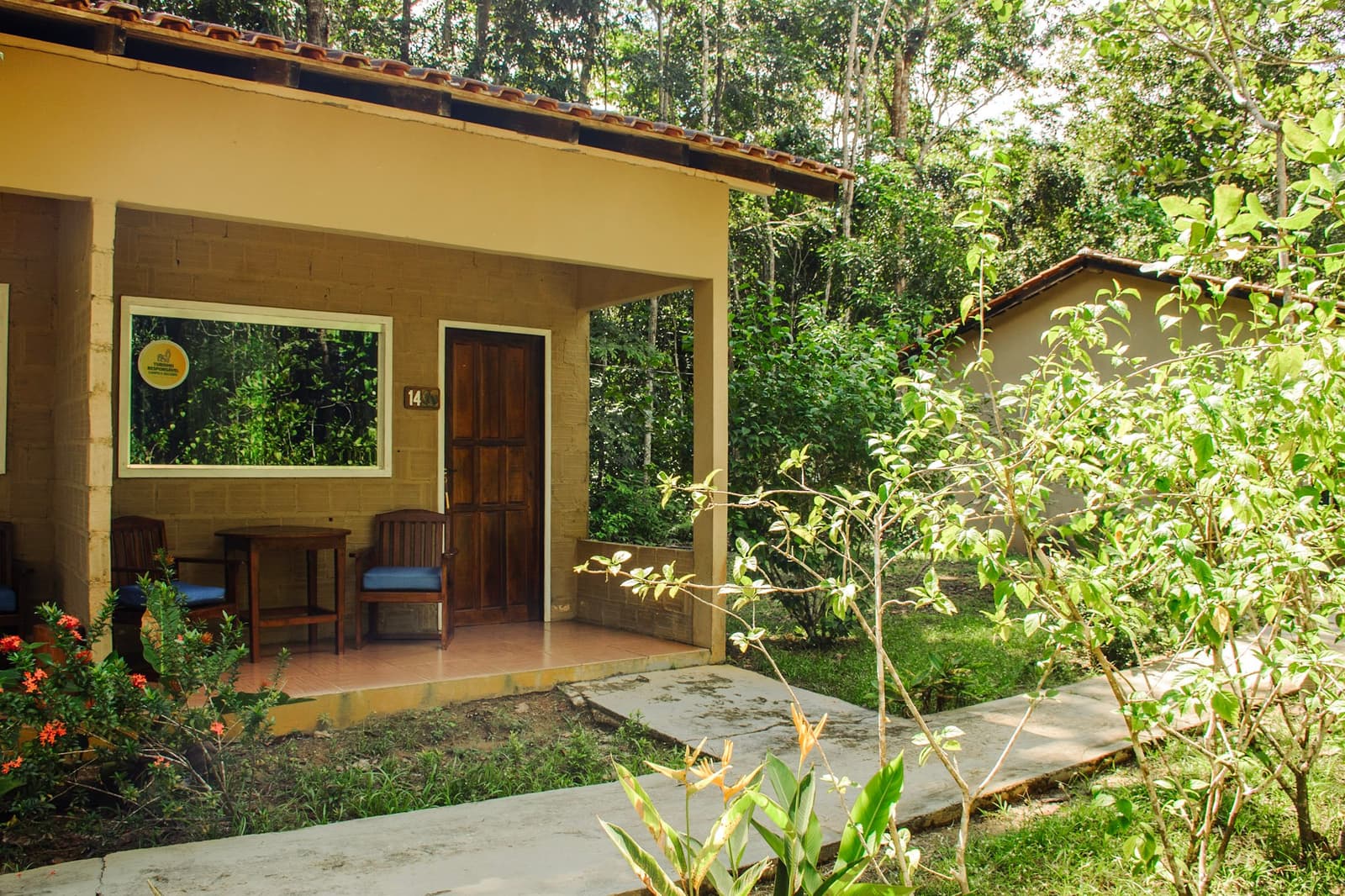 Room 14 has more trees than neighbours, which is exactly the point. Exterior of a numbered jungle bungalow with a covered veranda, wooden chairs, and lush forest backdrop, part of an amazon and pantanal tour lodge stay