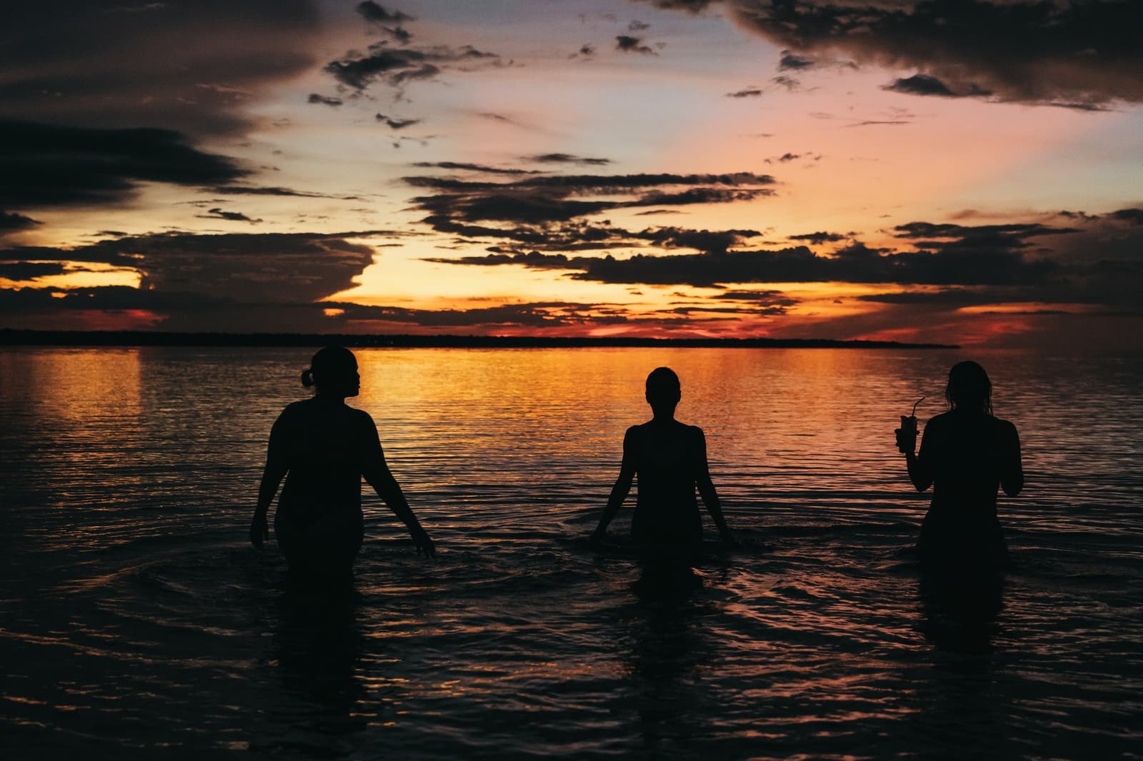 Warmes Wasser, leuchtender Himmel, ein Getränk in der Hand. Drei Personen als Silhouetten stehen hüfttief in einem breiten Fluss bei einem Luxus-Amazonas-Kreuzfahrt in Brasilien, während sich ein leuchtendes Orange-Rot des Sonnenuntergangs im ruhigen Wasser spiegelt